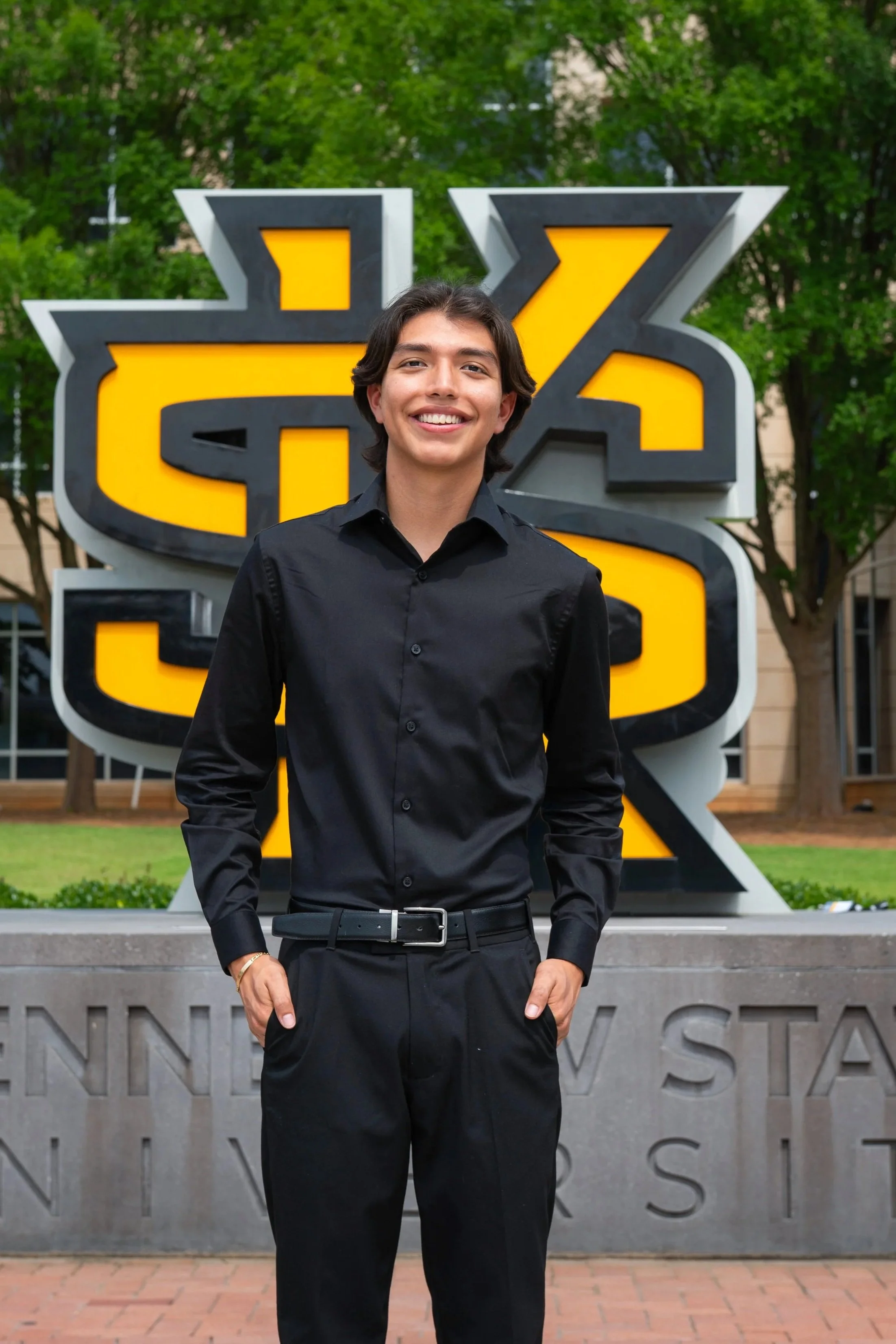 A woman in a black dress smiling and standing outdoors in front of a large yellow and black emblem, possibly at a university or institution.