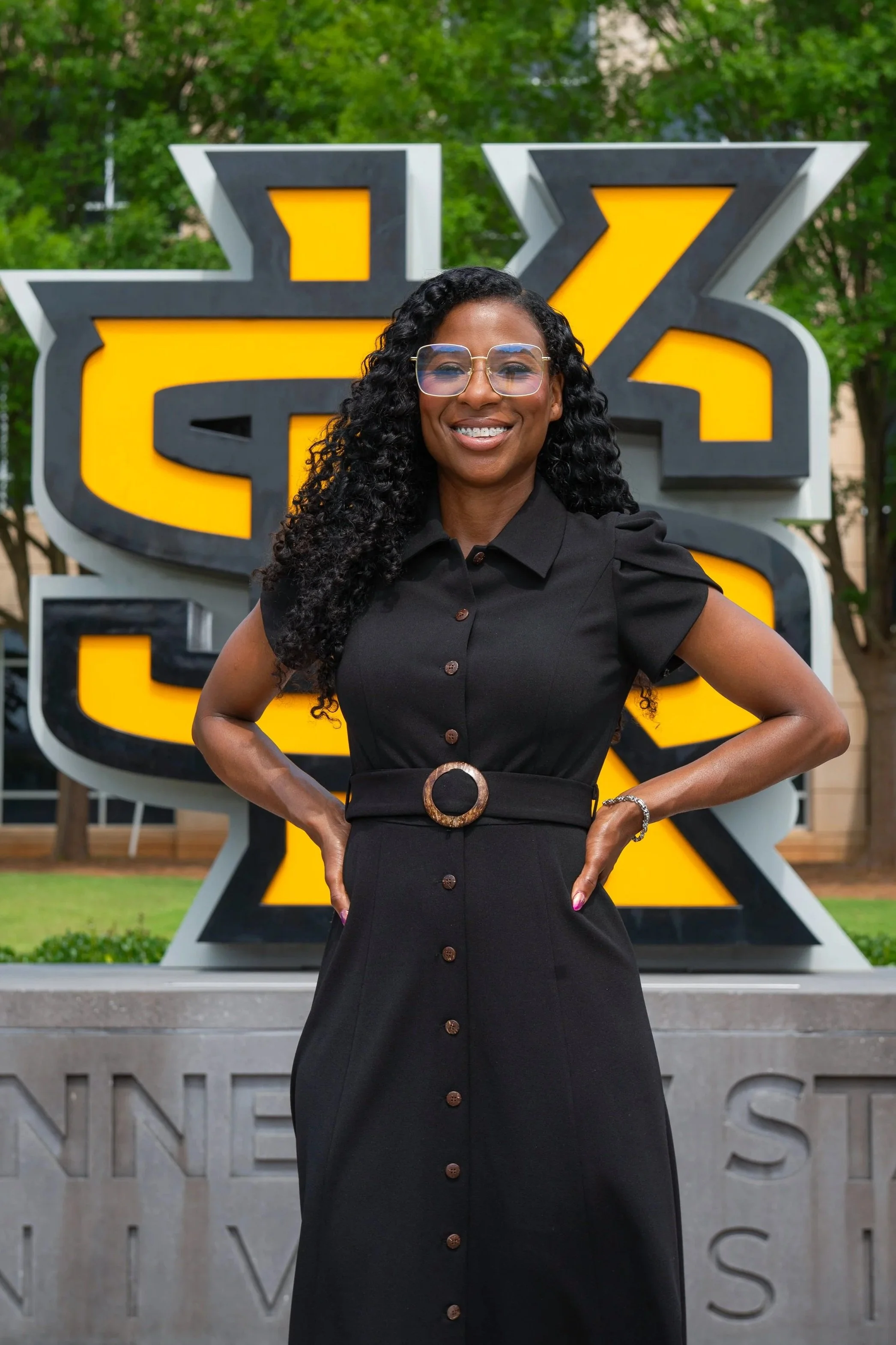 A woman in a white cardigan and black outfit stands outdoors in front of a university sign with trees and a fence in the background.