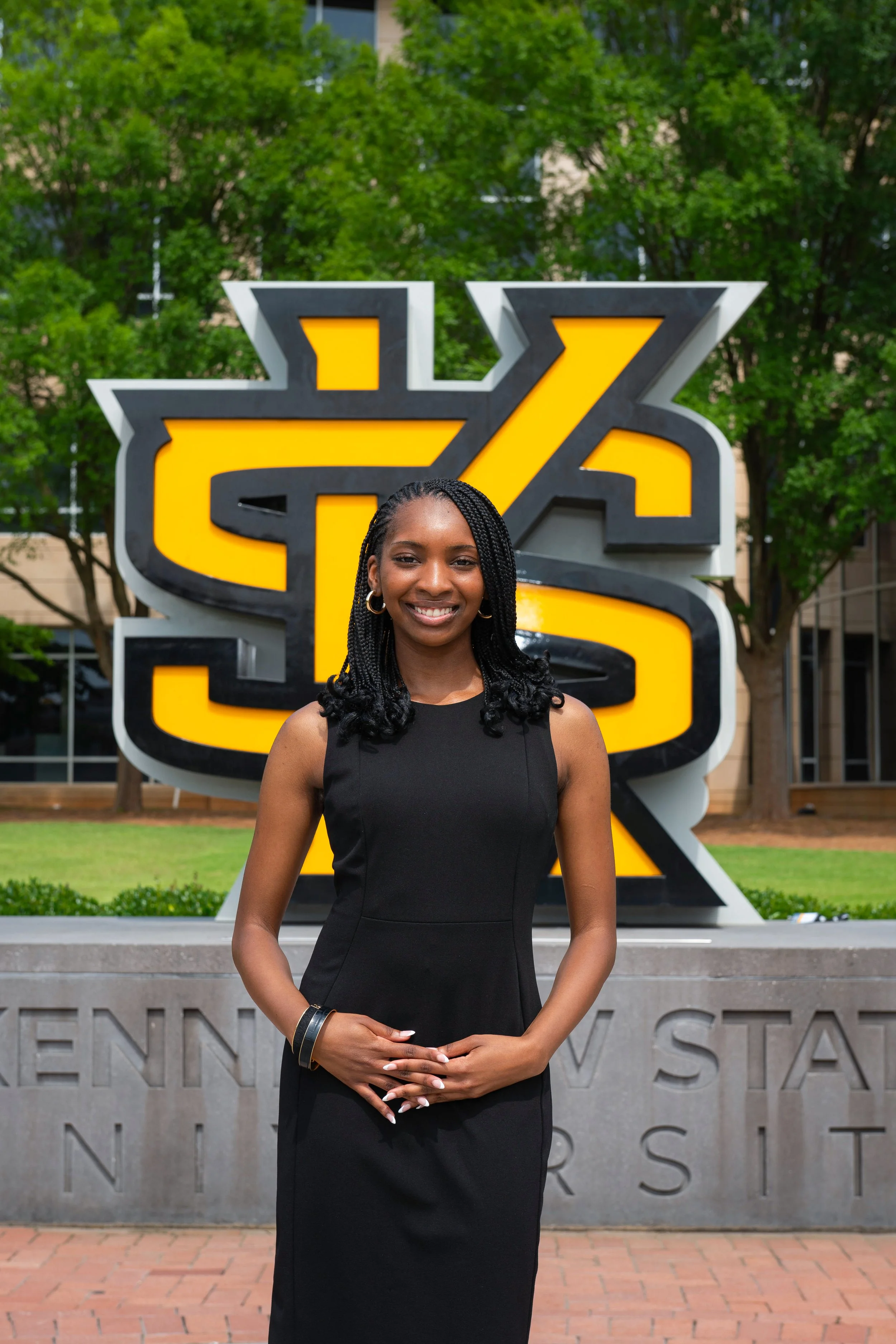 A smiling woman with glasses and braided hair standing outdoors in front of a large yellow and black sign with trees in the background.