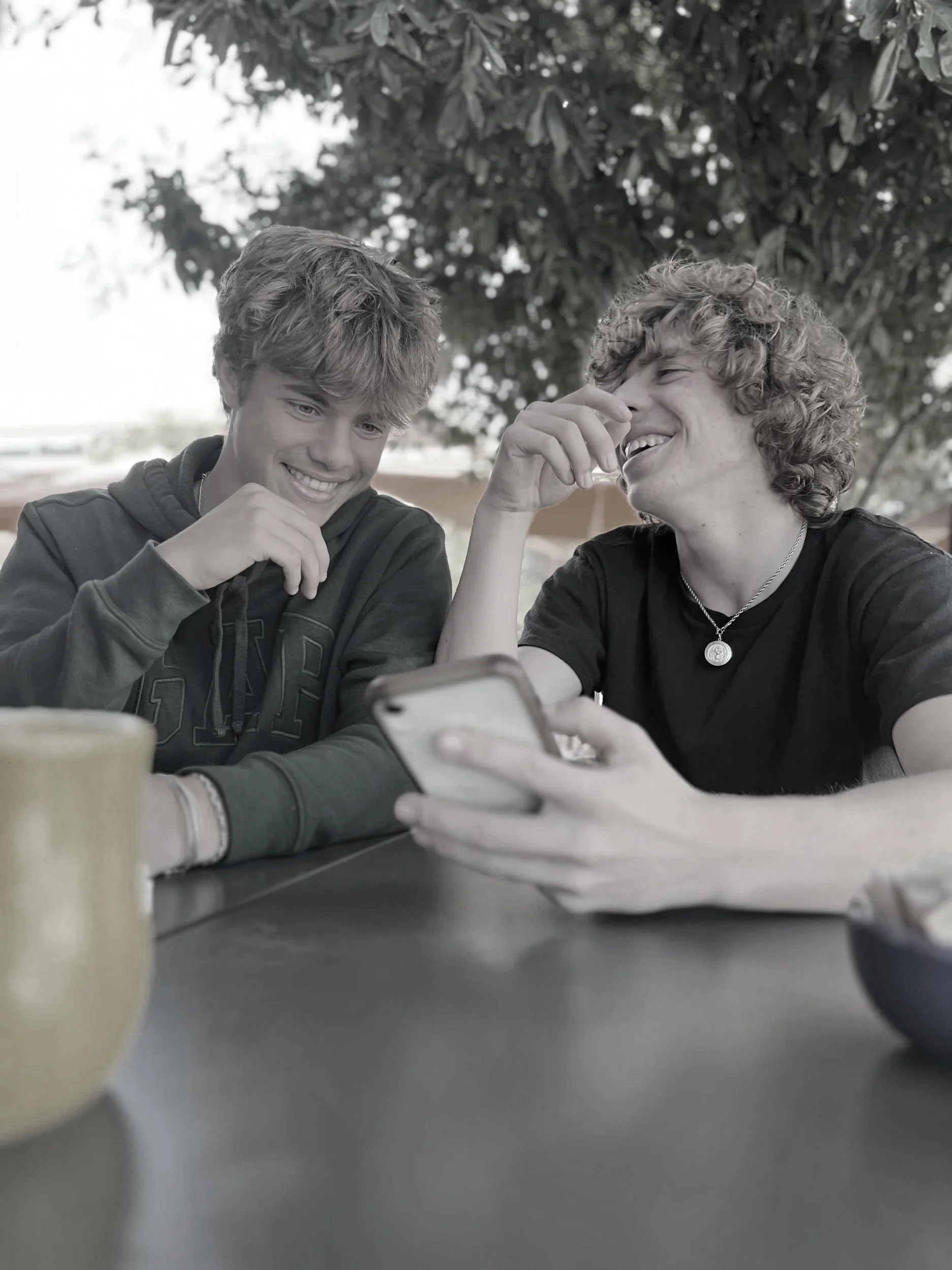 Two young teenagers sitting at a table outdoors, looking at a smartphone and laughing, with a tree and outdoor setting in background.