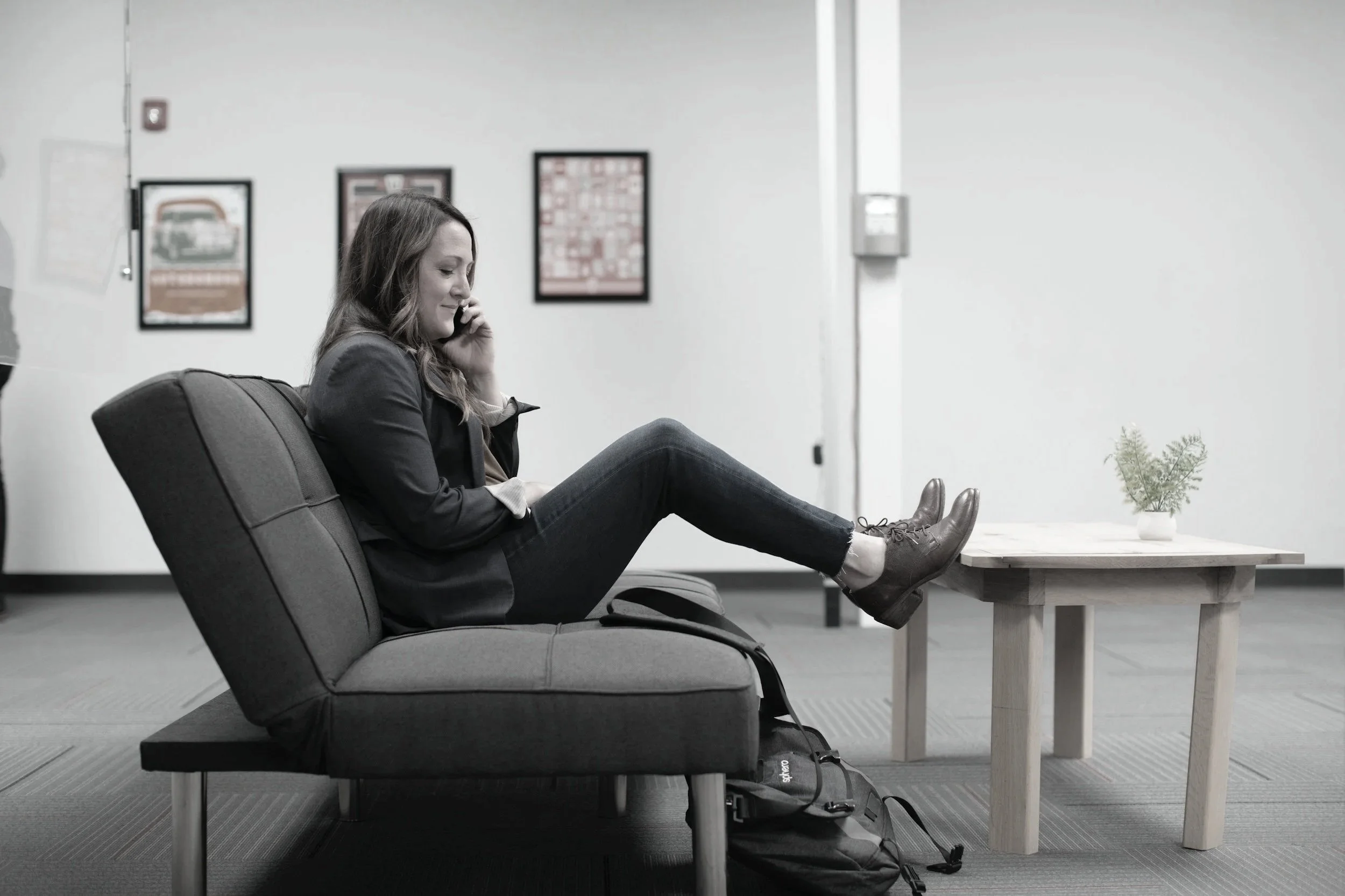 Teacher sitting on gray lounge chair talking on phone, with a small wooden table and potted plant nearby, in an office or waiting area.