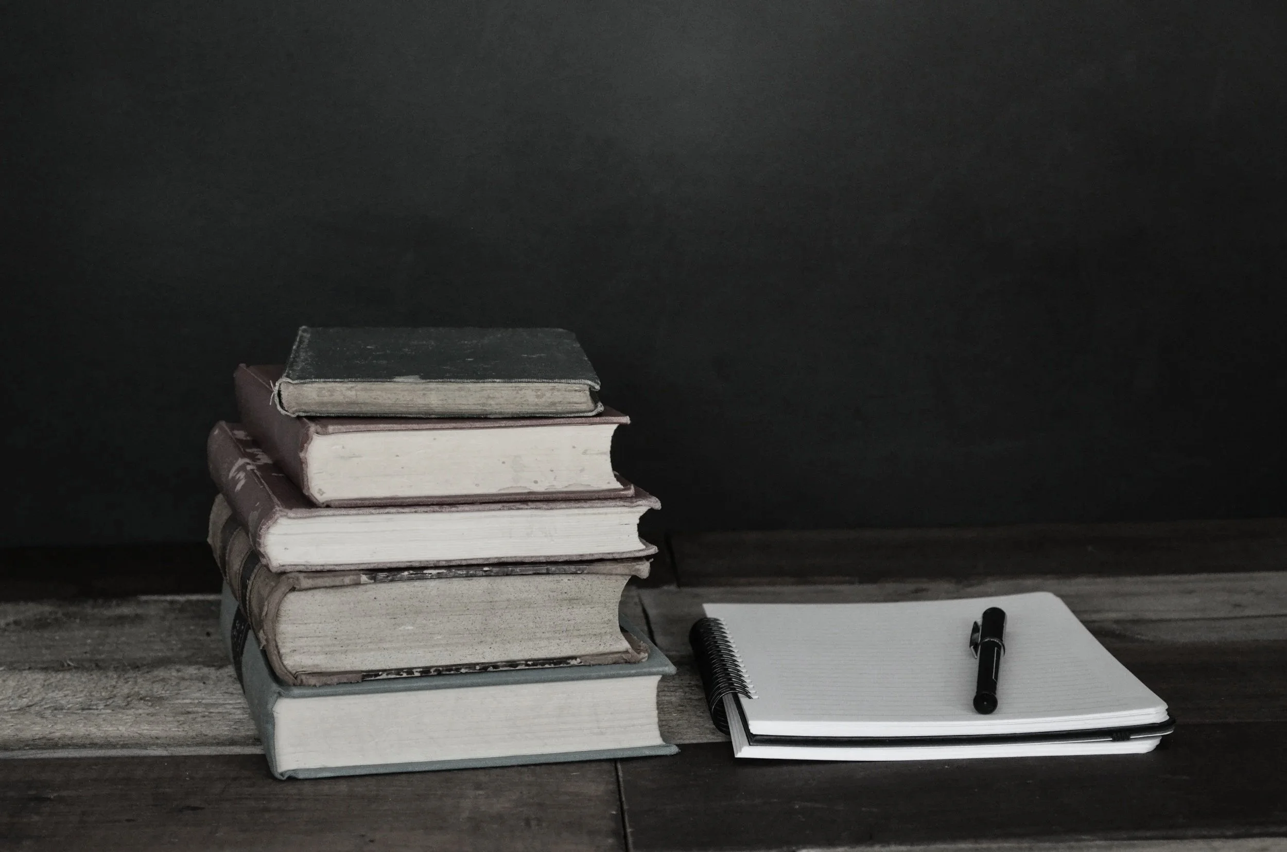 A stack of six old, worn books with a notepad and a pen on a wooden surface against a dark background.