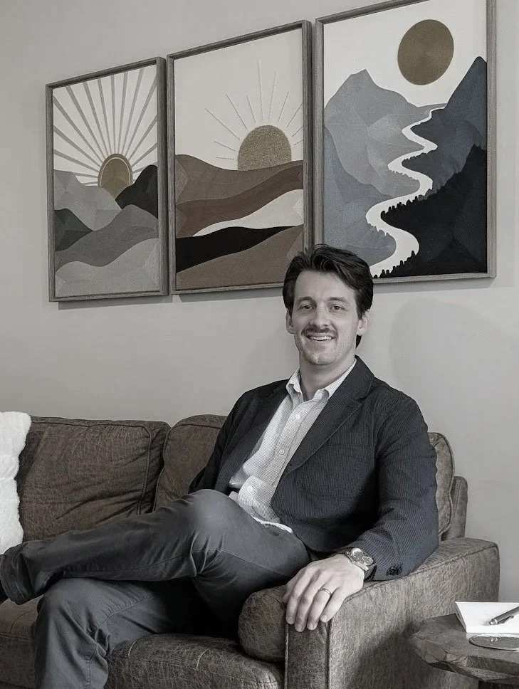 A man sitting on a brown sofa, smiling and wearing a suit, with three landscape art pieces depicting mountains, a river, and the sun hanging on the wall behind him.