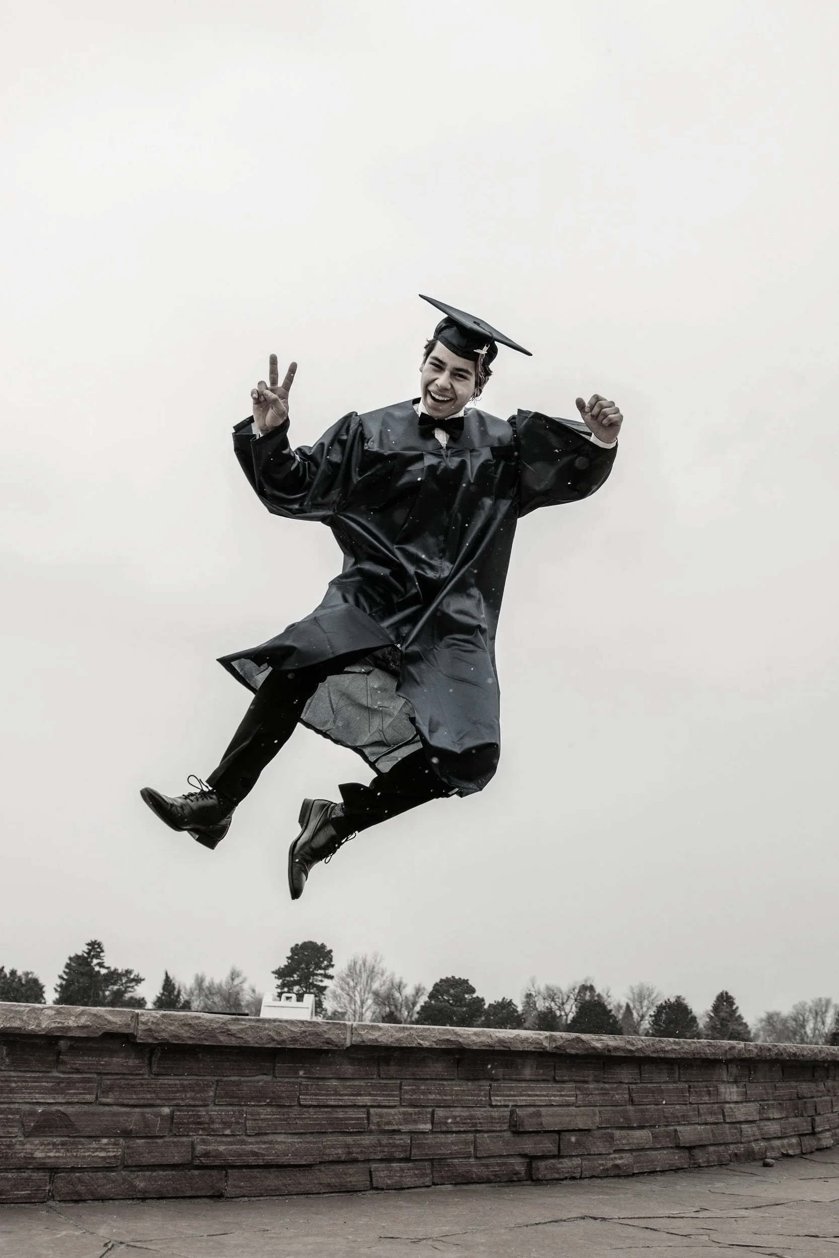 Young student in a graduation cap and gown jumpingly smiles, holding up a peace sign.