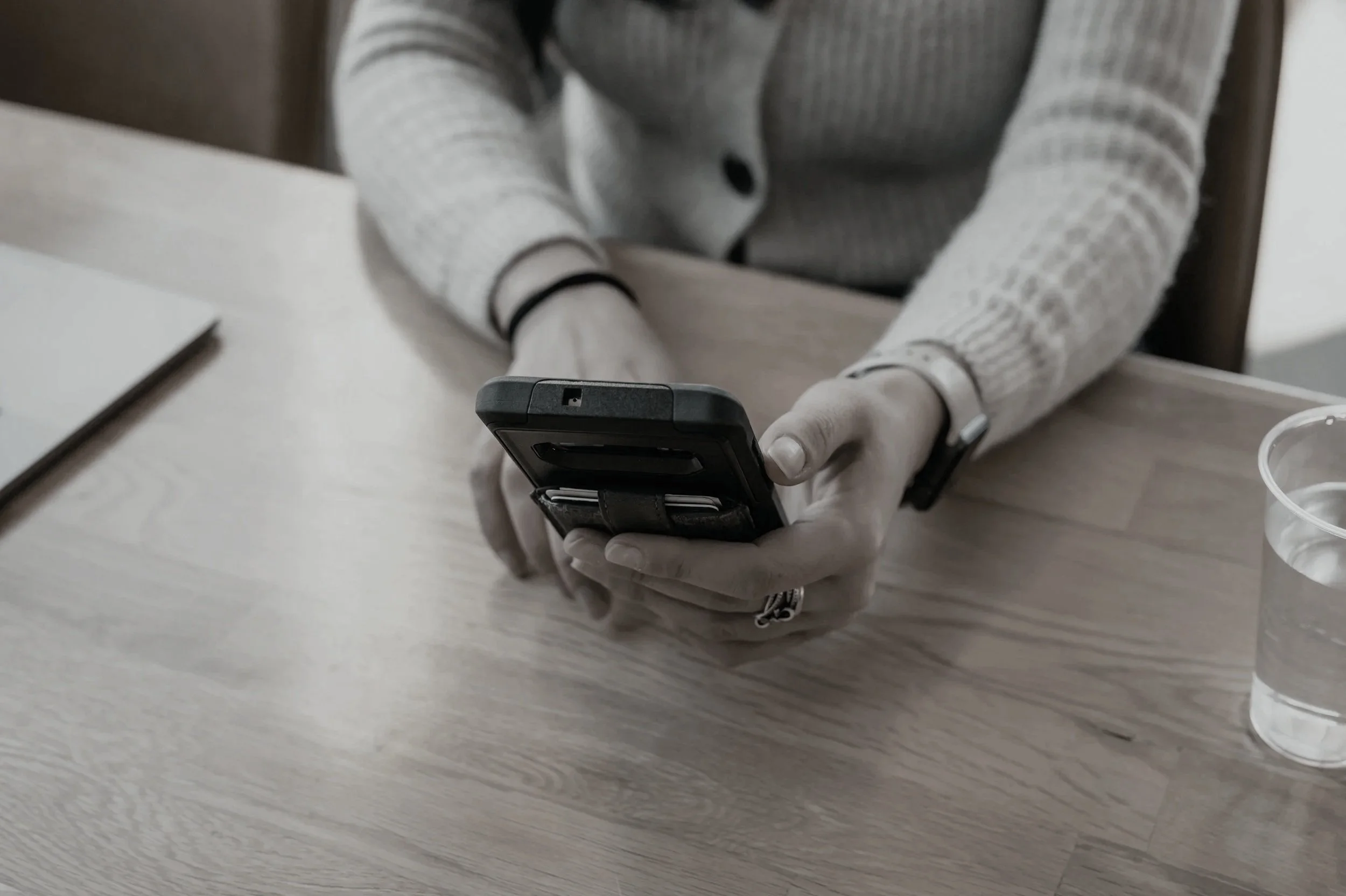Teacher in a sweater sitting at a wooden table, holding a smartphone in both hands, with a glass of water and a closed notebook on the table.