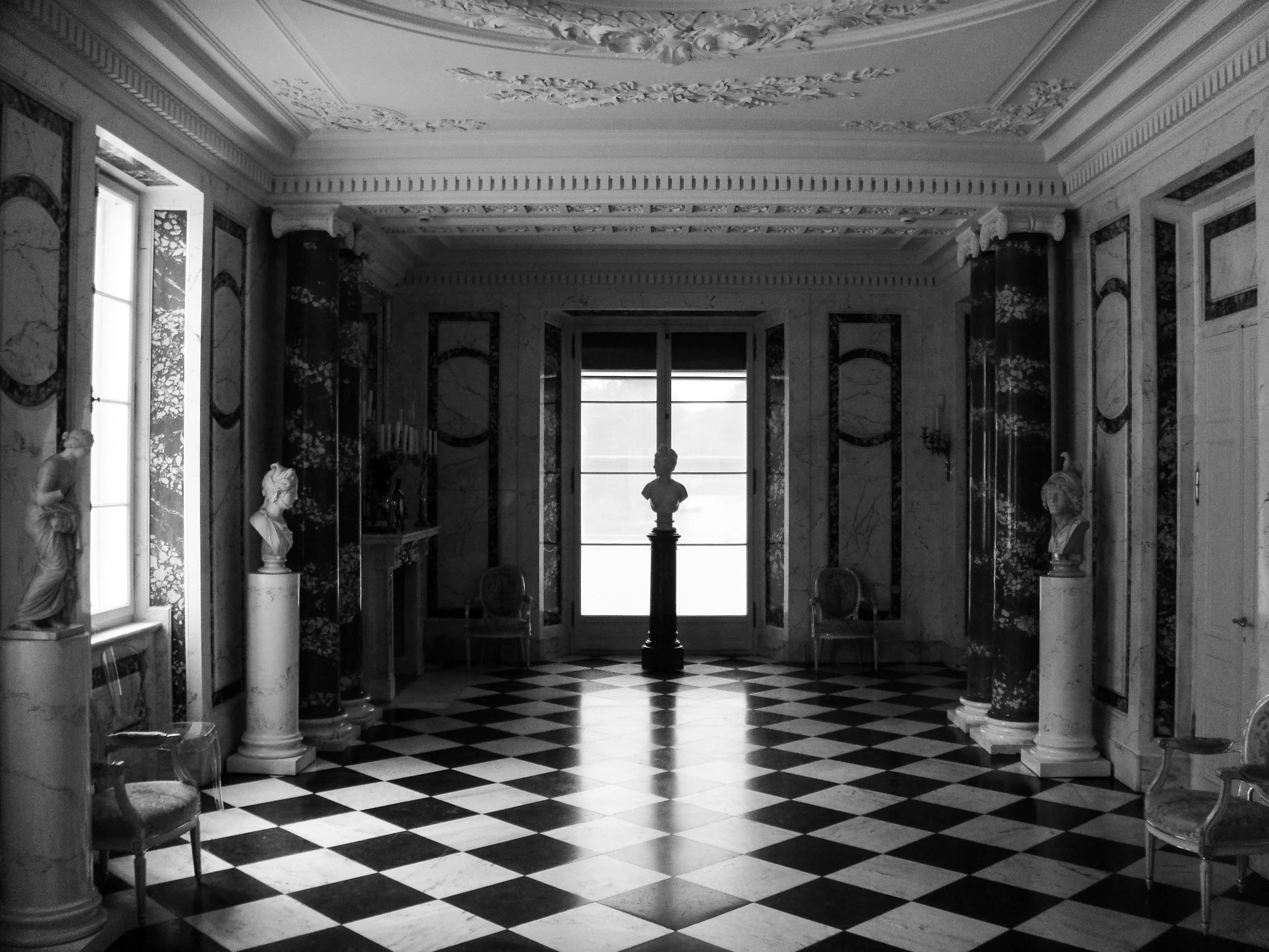 Black and white interior of a luxurious room with marble statues, pillars, high ceilings, and a checkered floor, featuring a window with a bust sculpture on a pedestal in front of it.