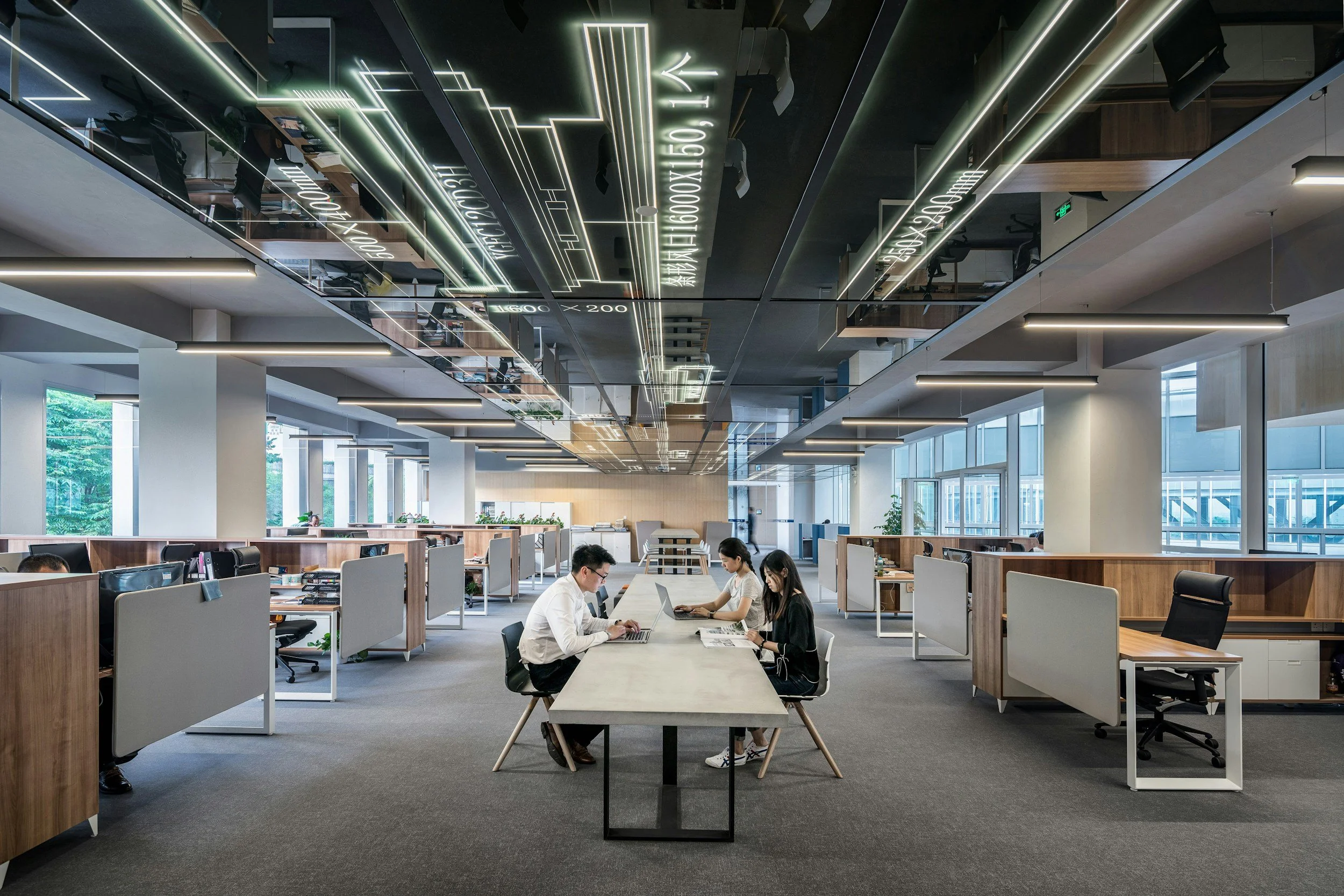 Modern open-plan office with three employees working at a long table, surrounded by cubicles, large windows, and ceiling lights.