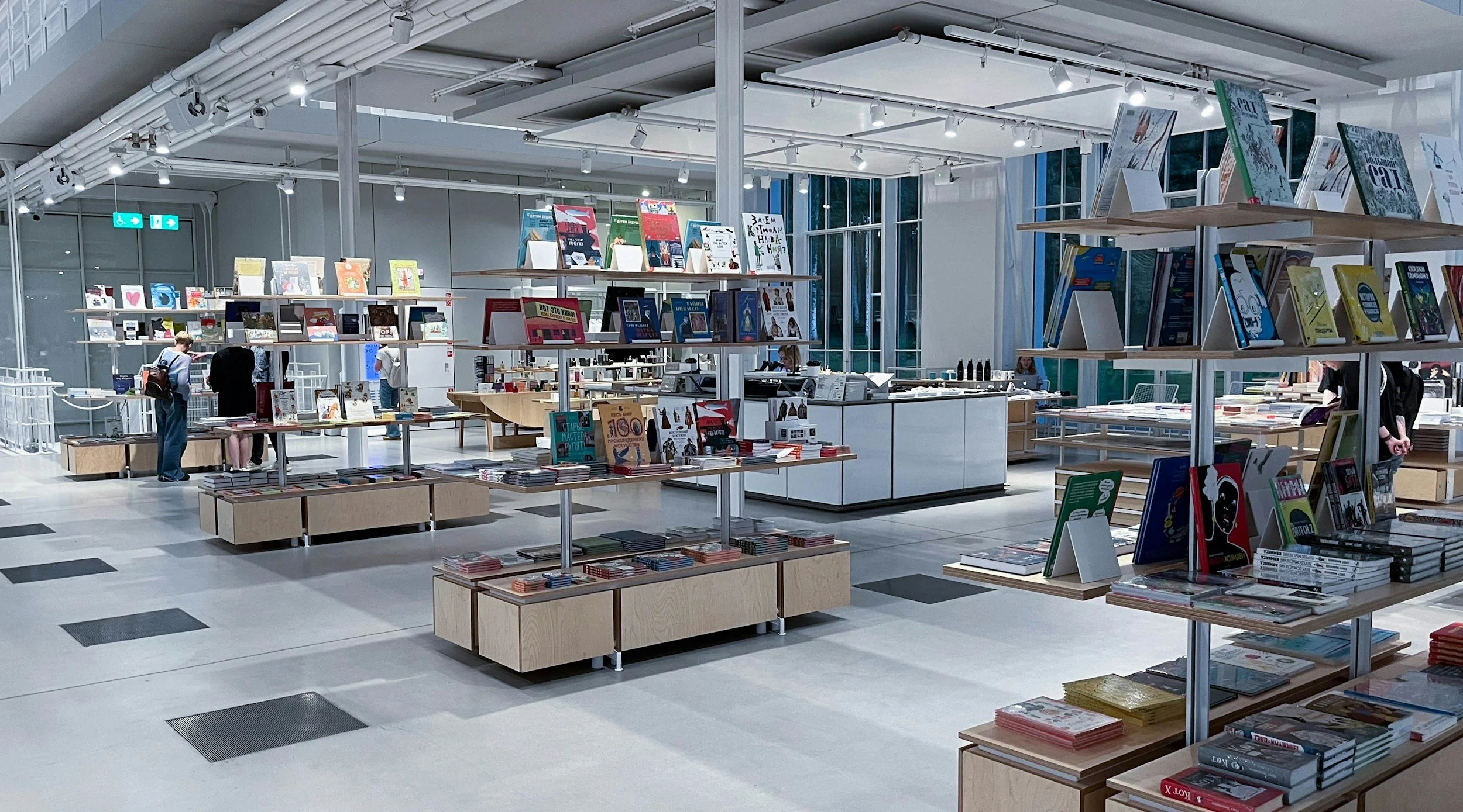 Inside a modern bookstore with books displayed on wooden tables and white display shelves, illuminated by ceiling track lighting, with a few customers browsing.