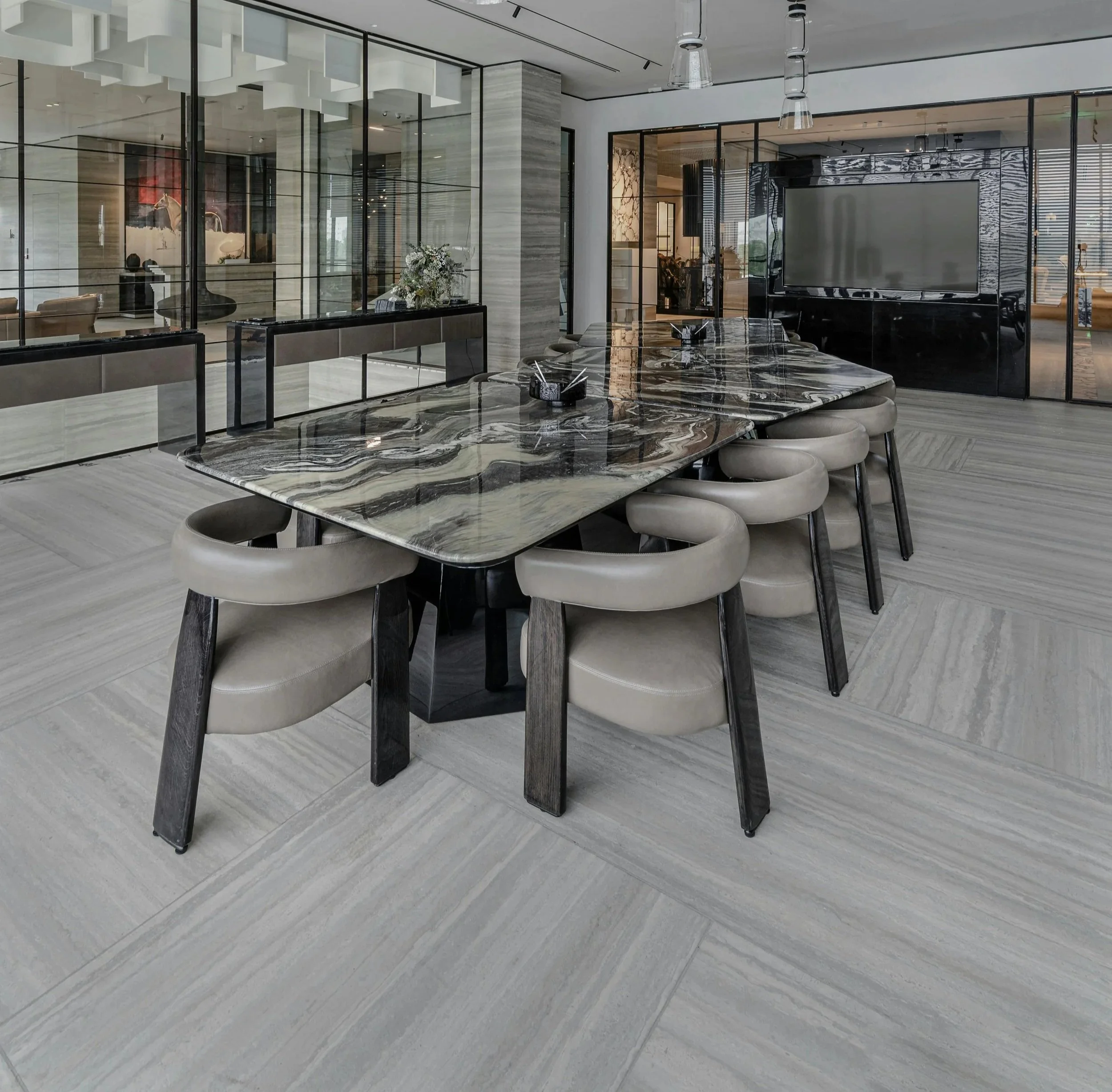 Modern conference room with a large marble dining table, beige upholstered chairs, glass partitions, light grey tile flooring, and a large flat screen TV mounted on a black glass wall.