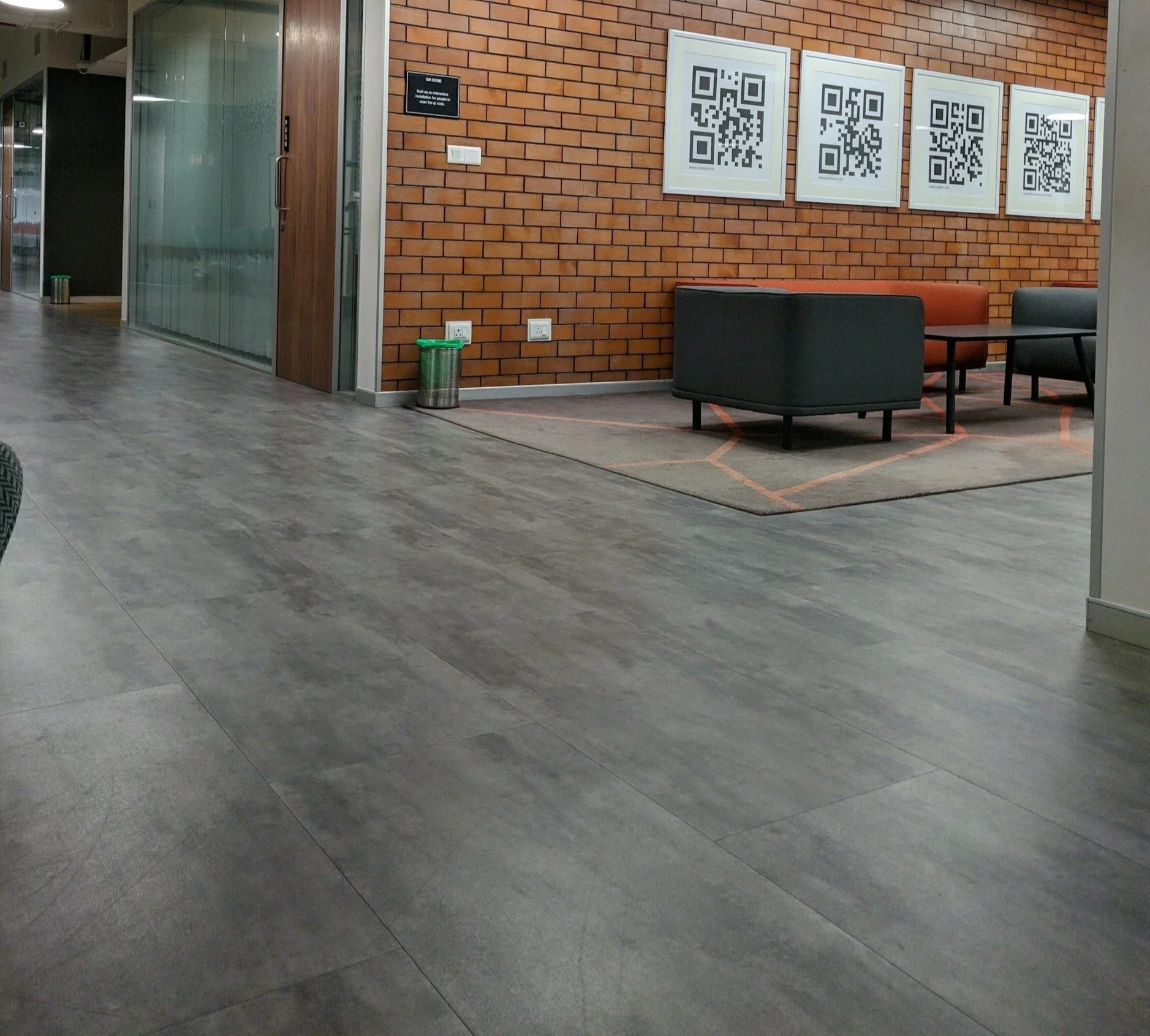 Modern office waiting area with brick wall, four framed QR code posters, black and orange armchairs, and a coffee table, illuminated by bright lighting.