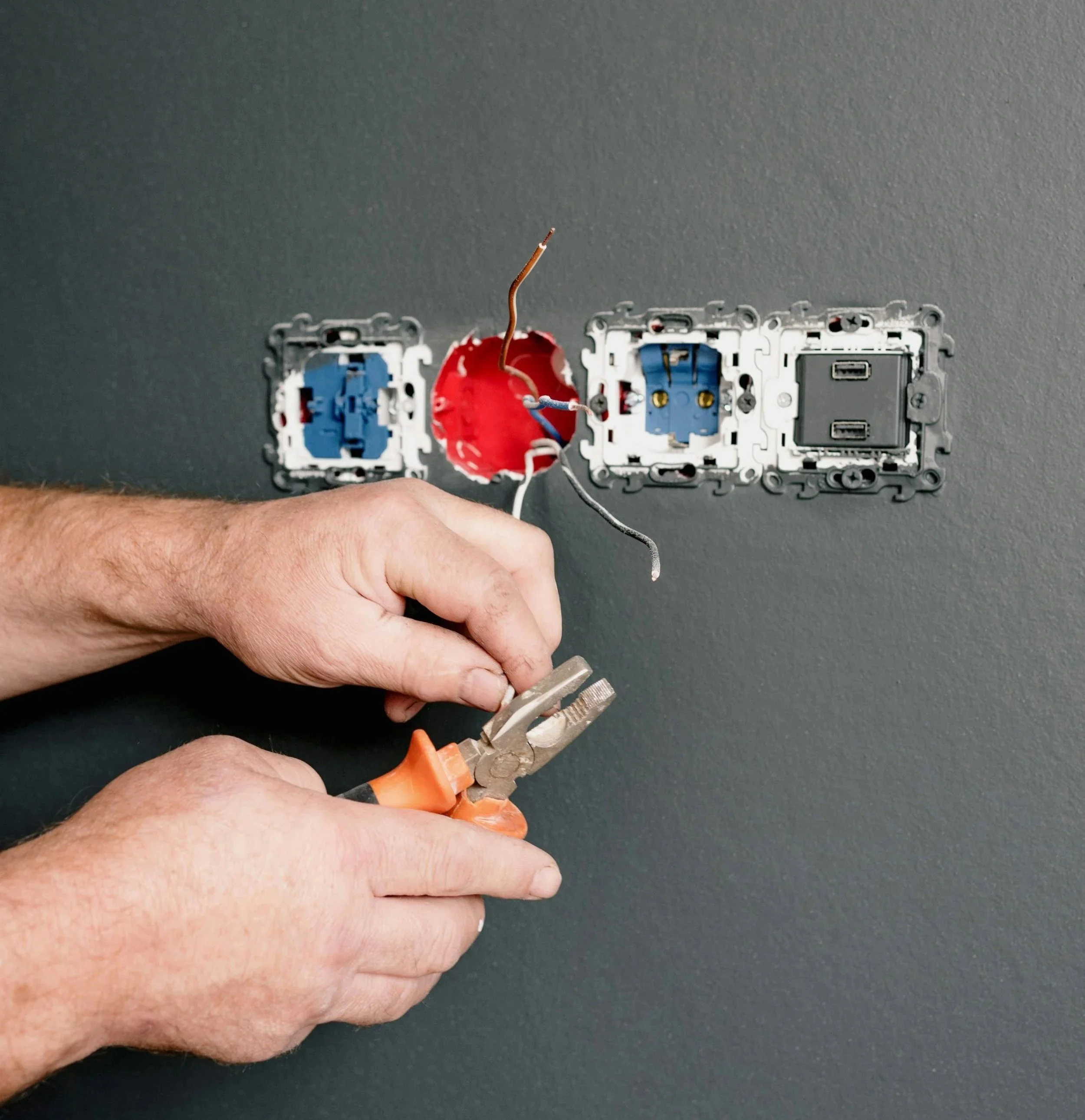 Hands working on electrical wiring behind a wall with three electrical outlets and a red junction box, using pliers.