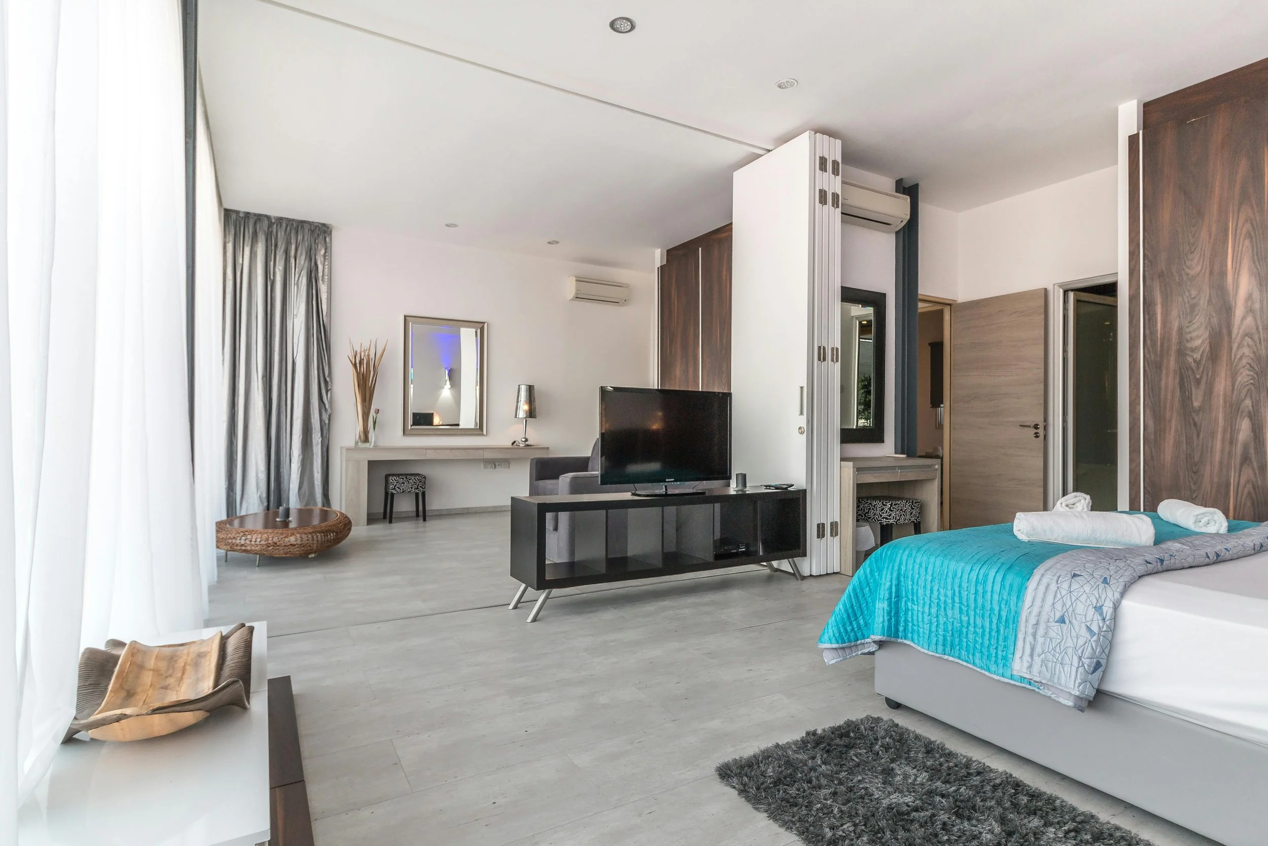 Modern hotel room with our bed, TV, sofa, small desk, curtains, and decorative elements.