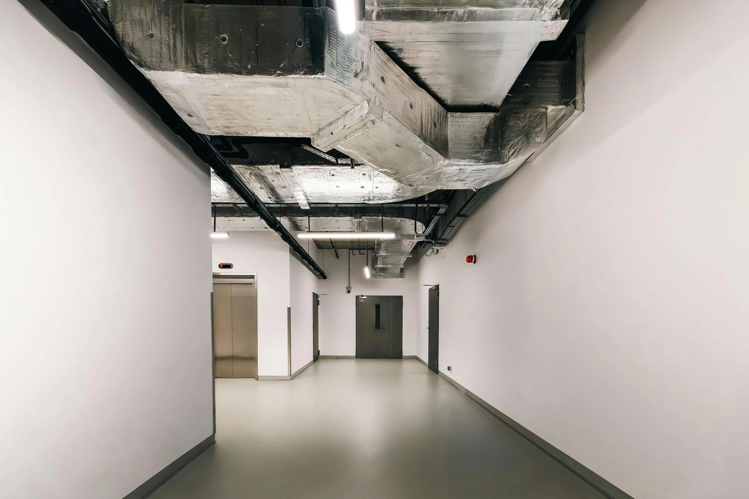 Empty hallway in a modern building with white walls, gray doors, and exposed silver and black ceiling ductwork.