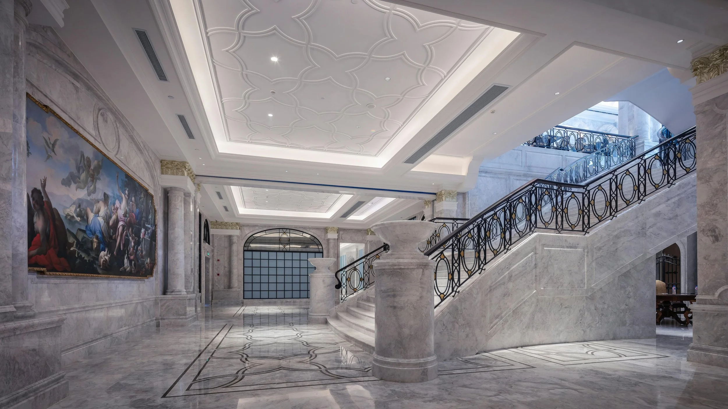 Elegant marble lobby with a staircase, classical artwork, and ornate ceiling details.