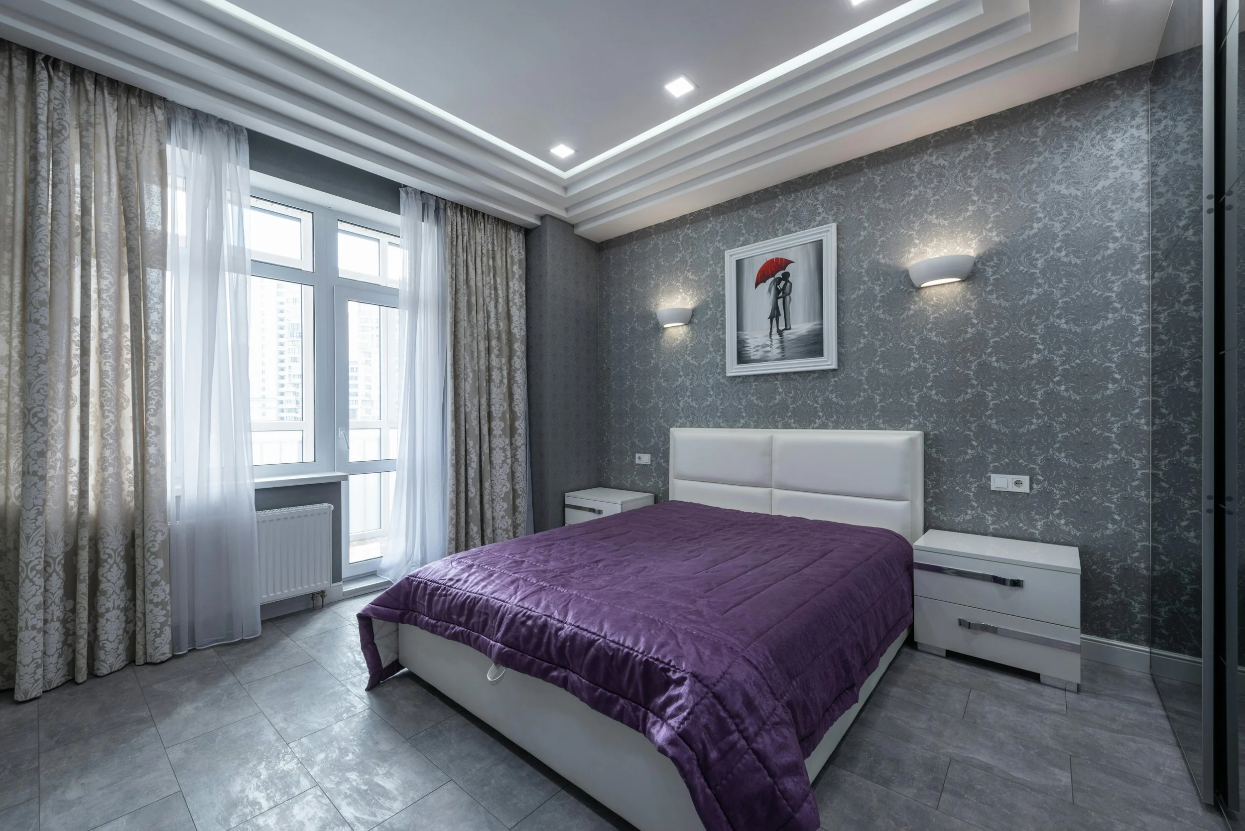 Modern bedroom with gray patterned wallpaper, white headboard, purple bedspread, two white nightstands, sheer and patterned curtains, framed artwork of a couple under an umbrella, and multi-layered ceiling lighting.