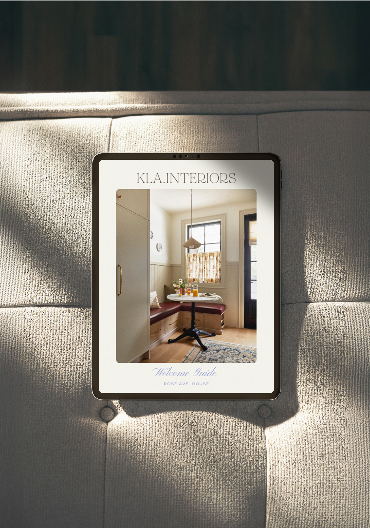 Digital tablet displaying a interior design company brochure titled 'KLA. INTERIORS' with a picture of a cozy kitchen nook and a welcome guide text, placed on a gray upholstered surface.