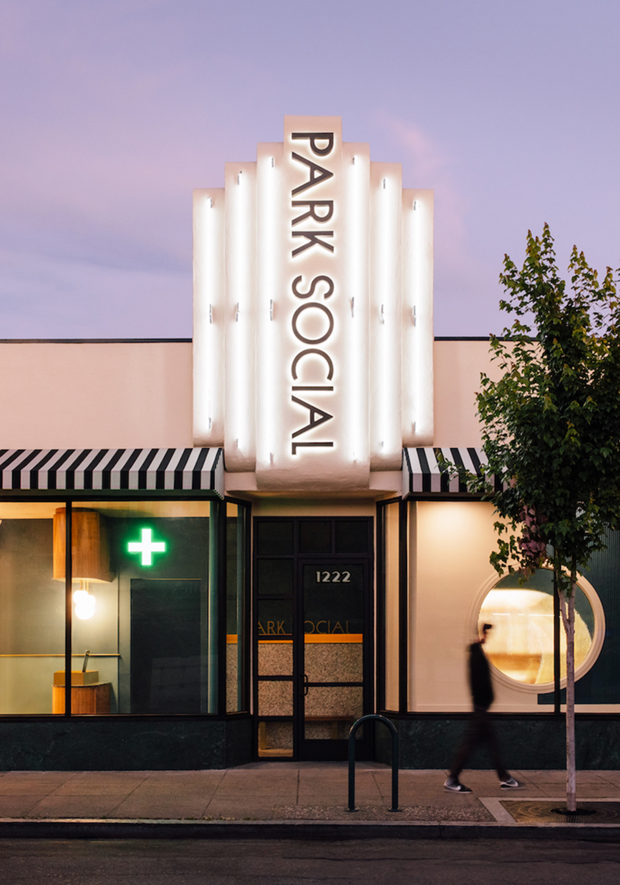 The front of a building with a large vertical sign reading 'PARK SOCIAL.' The building has a black and white striped awning, a green pharmacy cross sign, and a glass entrance door with the number 1222 above it. There is a person blurred walking past, and a small tree on the right side.