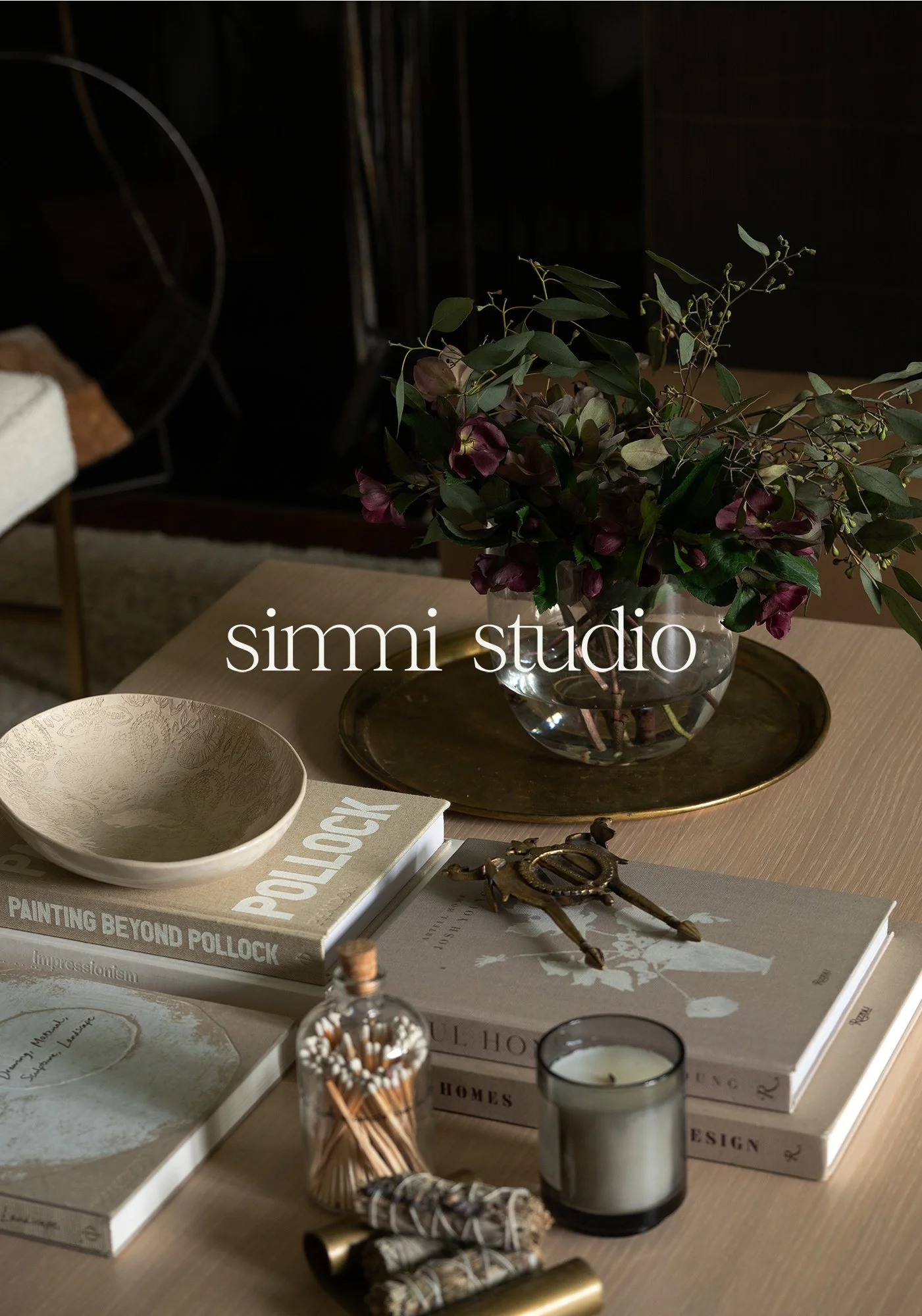 A bouquet of mixed flowers and foliage in a glass vase on a wooden table, surrounded by books, candles, and decorative items, with a dark background.