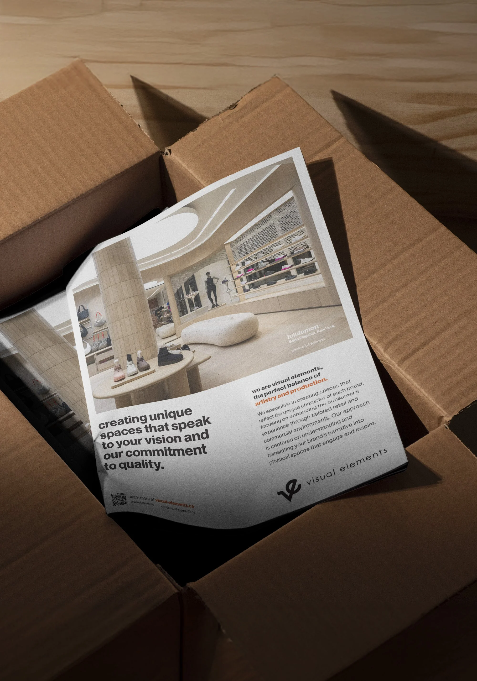 A printed flyer inside an open cardboard box promoting a company called Visual Elements, specializing in creating unique retail spaces. The flyer features an interior design image with modern decor, shoes displayed on racks, and a person standing in the space.