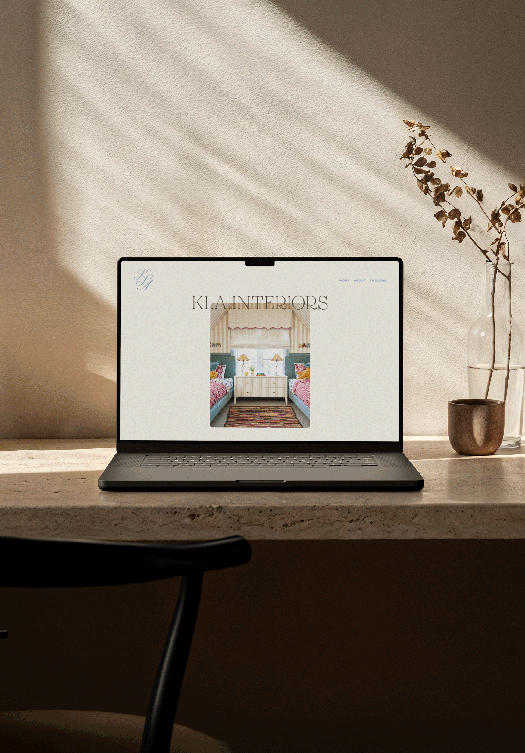 A laptop on a wooden desk displaying a webpage titled "KLA INTERIORS" with a bedroom interior photo. A glass vase with a dried branch is on the right side of the desk.
