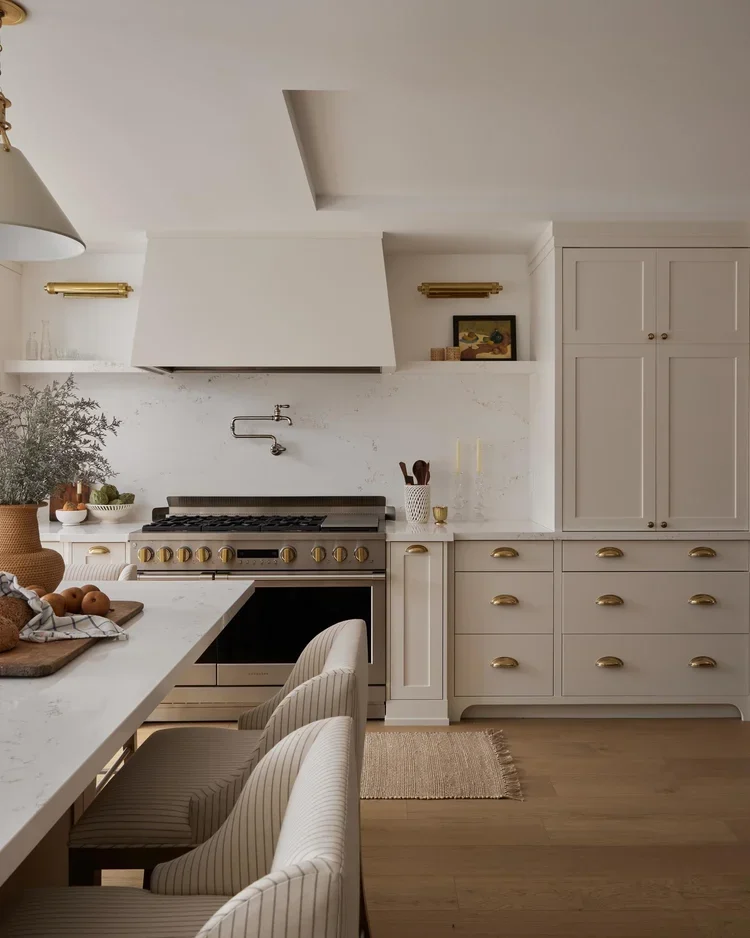 Modern kitchen with white cabinetry, gold hardware, and a large stove with a white marble countertop island. There are three striped chairs at the island, and decorative items like a plant, bowls, utensils, and candles are visible.