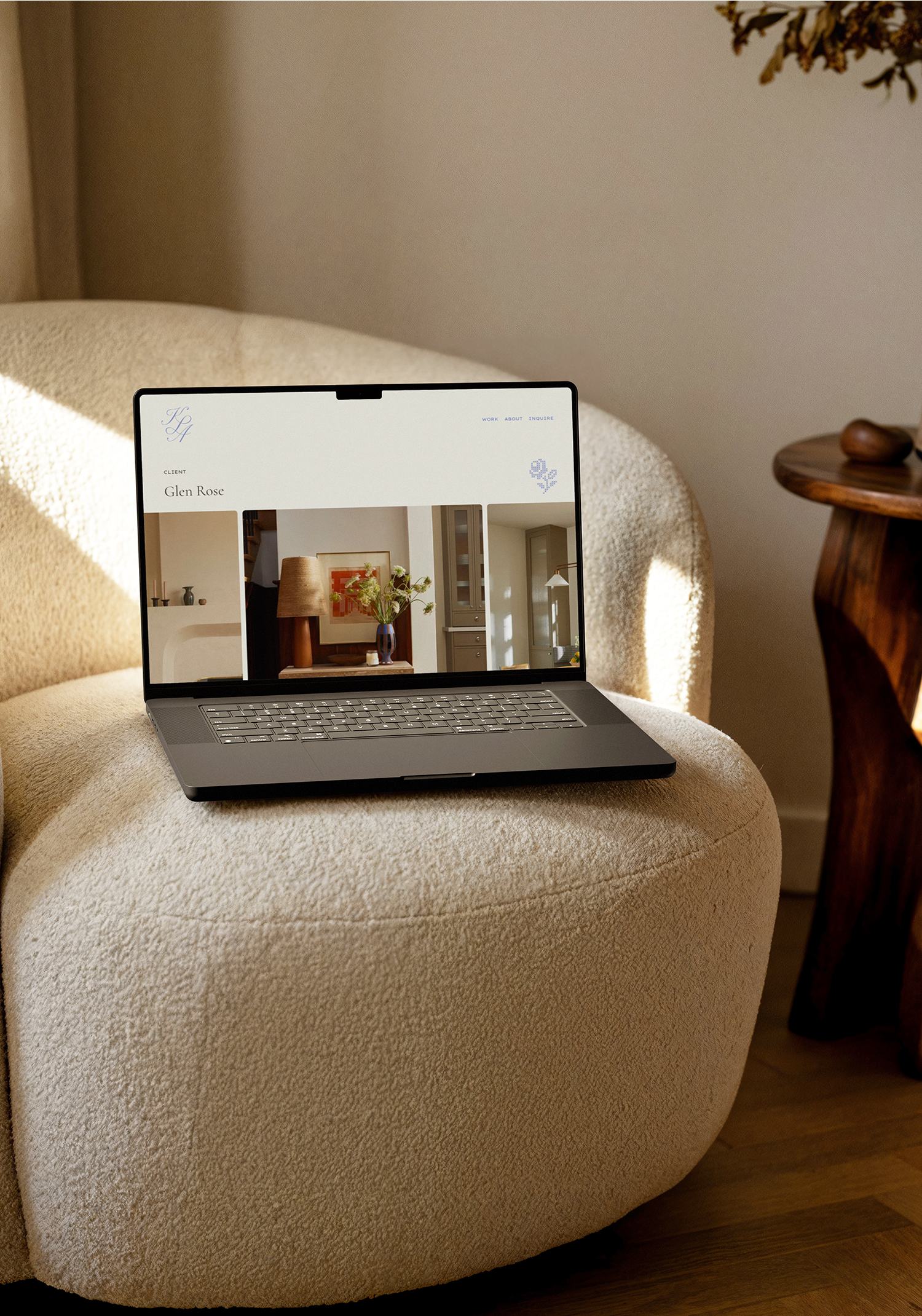 A laptop is placed on a plush beige armchair with a textured fabric. The laptop screen shows a website featuring interior decor images. To the right of the armchair, there is a wooden side table with decorative items.