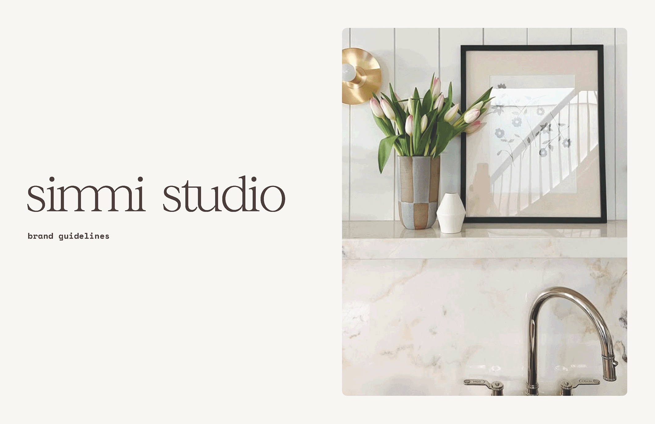 Interior of a bathroom with a marble countertop, a mirror, a vase of pink and white tulips, a white vase, a gold wall light, and the edge of a bathtub with a shiny chrome faucet.