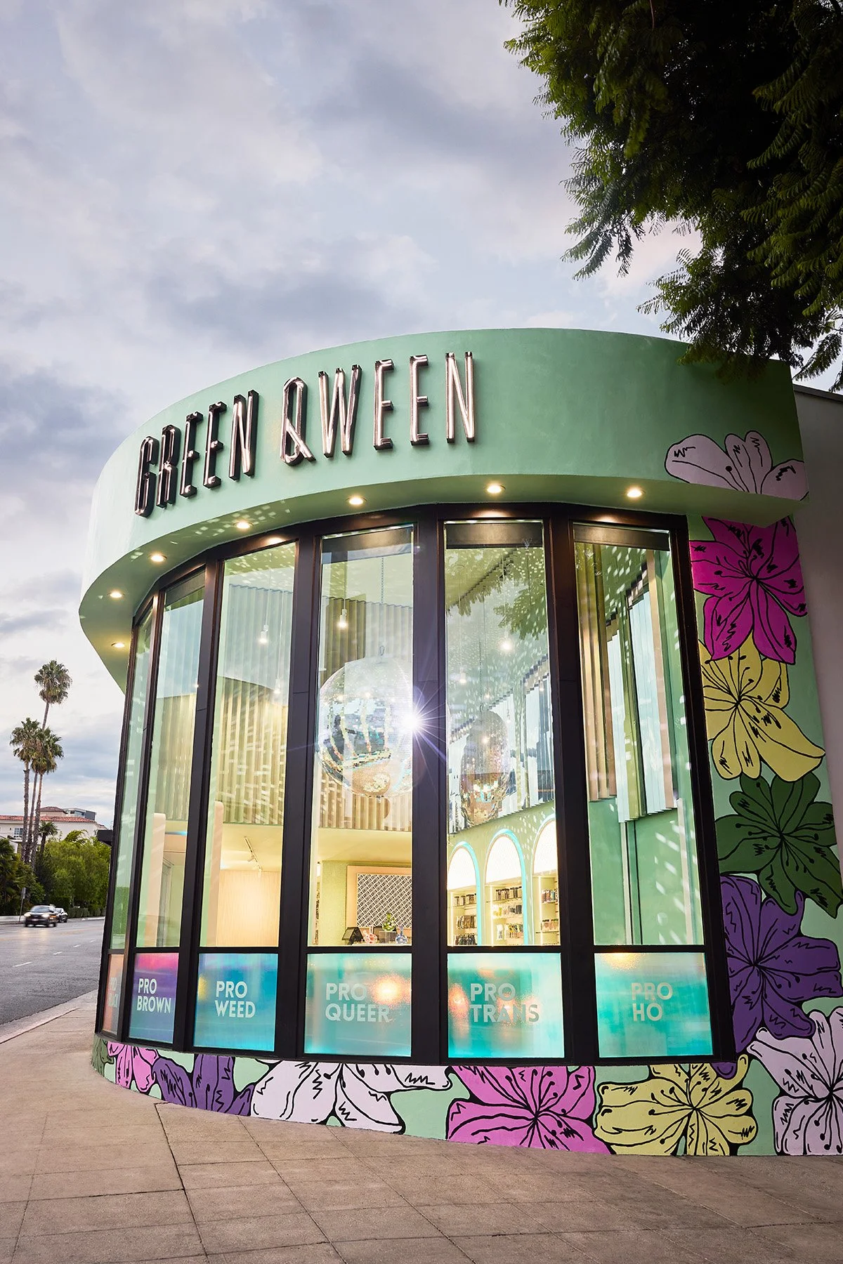 A storefront with the sign 'GHEEN &WEEN' on a rounded green facade, decorated with colorful floral artwork on the sides, large glass windows revealing the interior, and signs inside the windows reading 'PRO BROWN,' 'PRO WEED,' 'PRO QUEER,' 'PRO TRANS,' and 'PRO HO.'