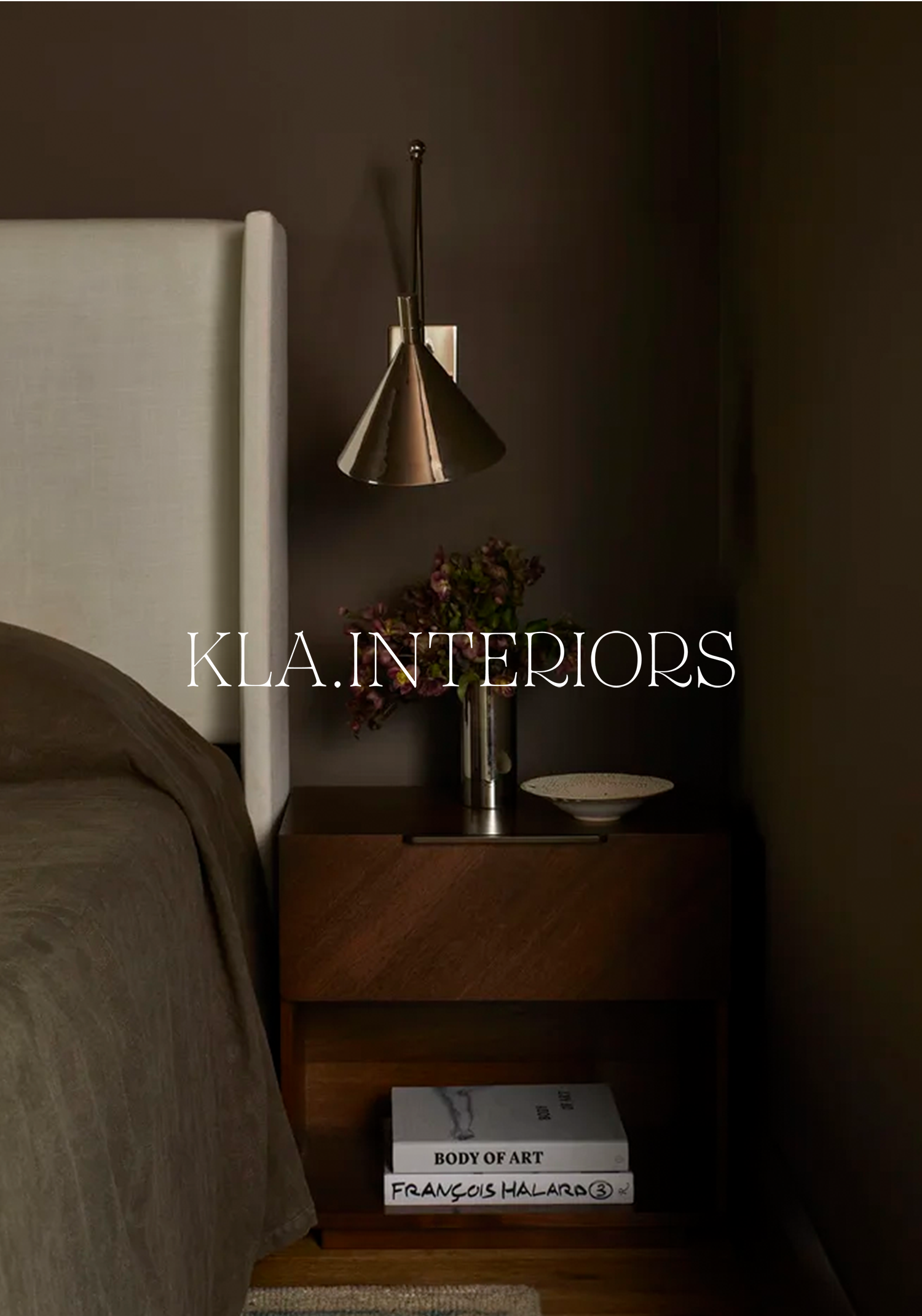 Nightstand with a silver wall-mounted lamp, a bowl, a plant, and books titled "Body of Art" and "Francis Halard" in a bedroom with dark walls and a bed with beige bedding.