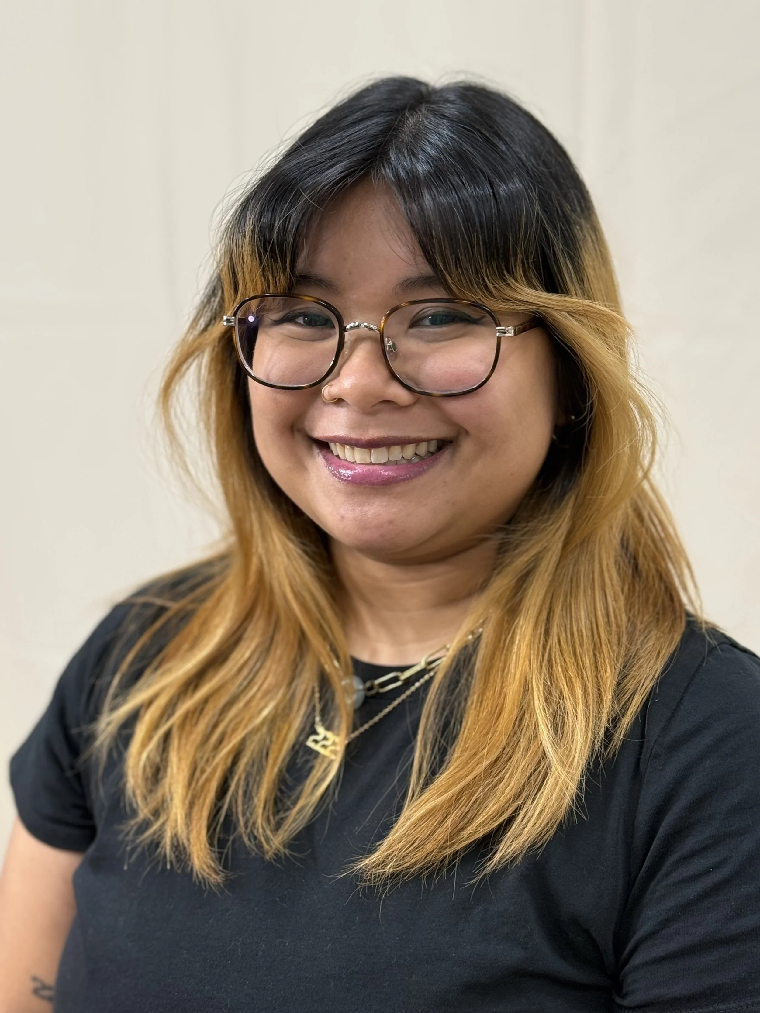 A smiling woman with shoulder-length black and blonde hair, wearing round glasses, a black shirt, and gold jewelry, standing against a plain light-colored wall.