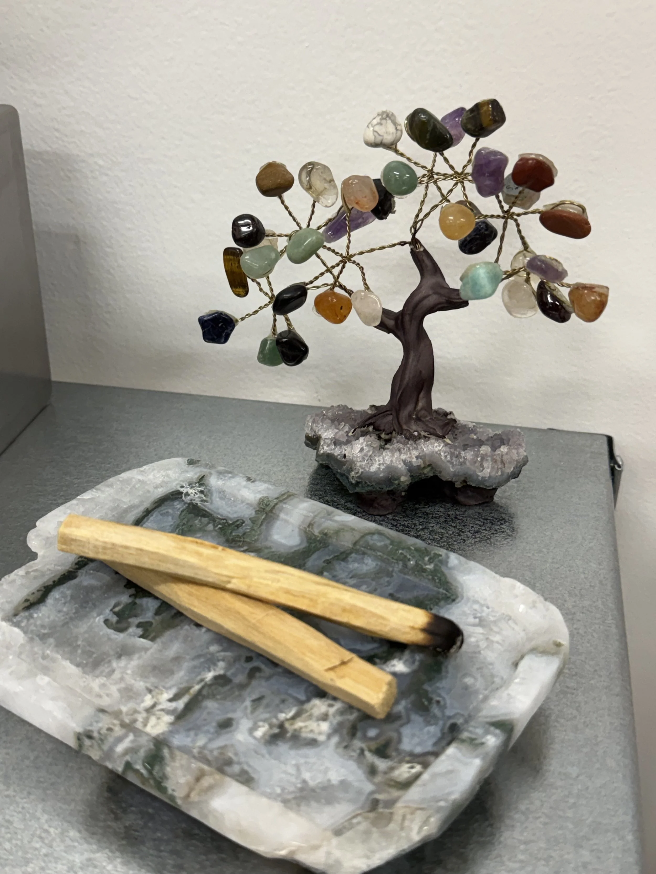 Decorative gemstone tree with multicolored stones and wire branches, placed on a rocks base, next to wooden sticks on a marble slab, on a gray table.