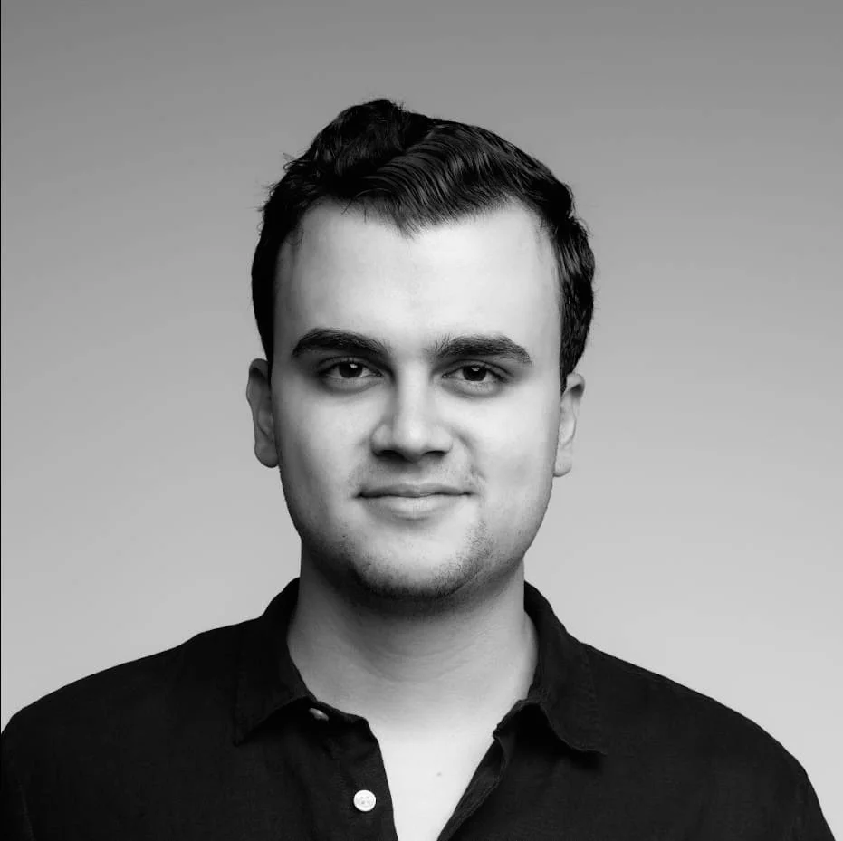 Black and white portrait of a young man with dark hair styled to the side, wearing a collared shirt, named Daniel Jaramillo, who is a managing partner at Raiz&Co, smiling slightly against a plain background.
