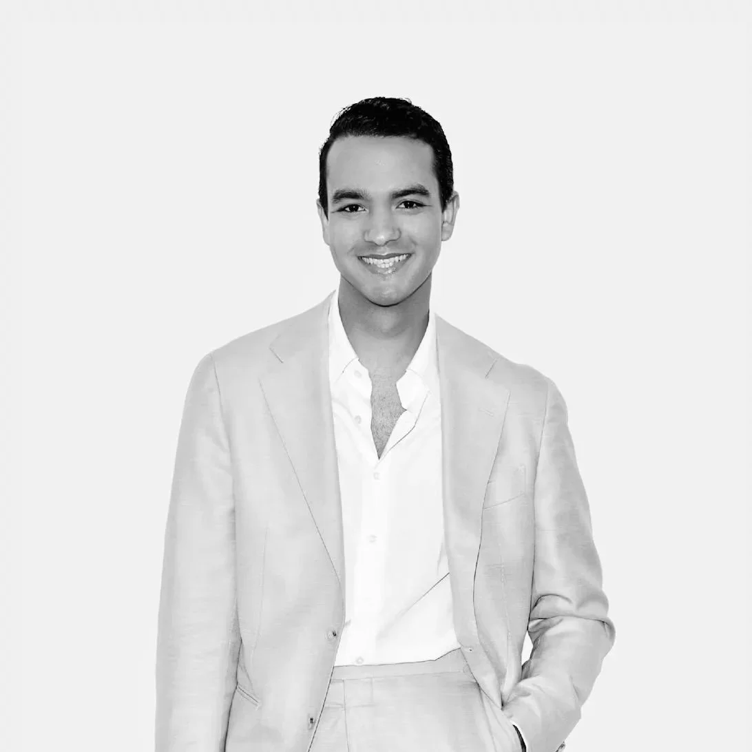 Black and white portrait of a young man named Randolhf Sanchez, who is the principal managing partner at Raiz&Co, in a light-colored suit and white shirt, smiling, with one hand in his pocket, standing against a plain background.