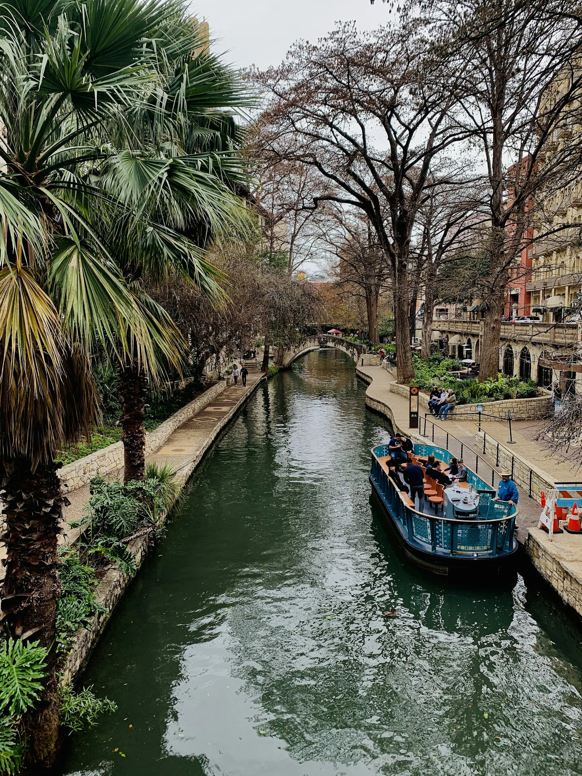 A canal scene with a boat carrying passengers and a DJ. Trees with no leaves line the canal, and there are buildings and pedestrians along the walkway.