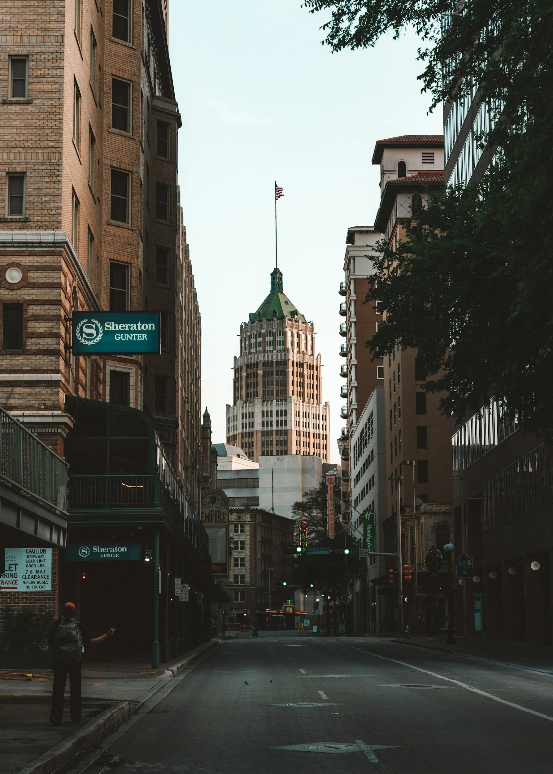 City street view with tall buildings, including a prominent skyscraper with a green cone-shaped roof and flag on top. There are hotel signs for Sheraton Gunter and Holiday Inn and a person with a backpack standing on the sidewalk.
