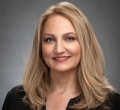 Portrait of a woman with blonde hair wearing a black blazer, smiling against a gray background.