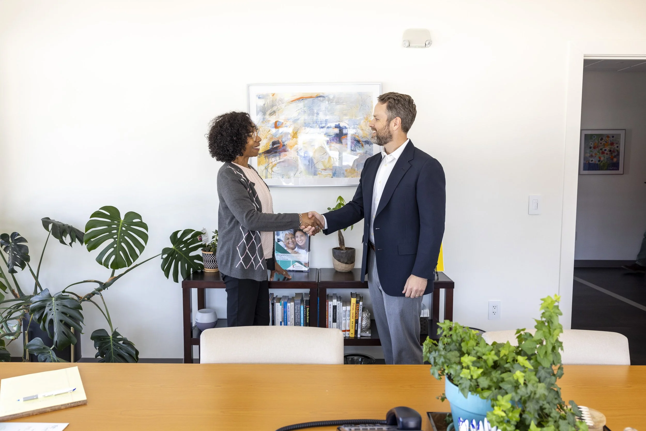 Two business professionals, a woman with curly hair and glasses and a man with a beard, shaking hands in an office.
