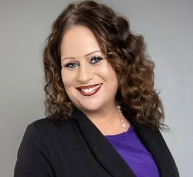 A woman with curly brown hair, wearing a black blazer over a purple top, smiles at the camera against a plain gray background.