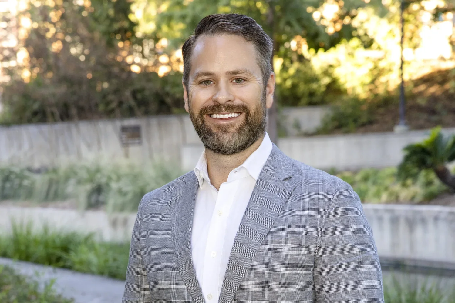 A man with gray hair and a beard, smiling, dressed in a light gray blazer and white shirt, standing outdoors in a park with trees and greenery in the background during sunset.