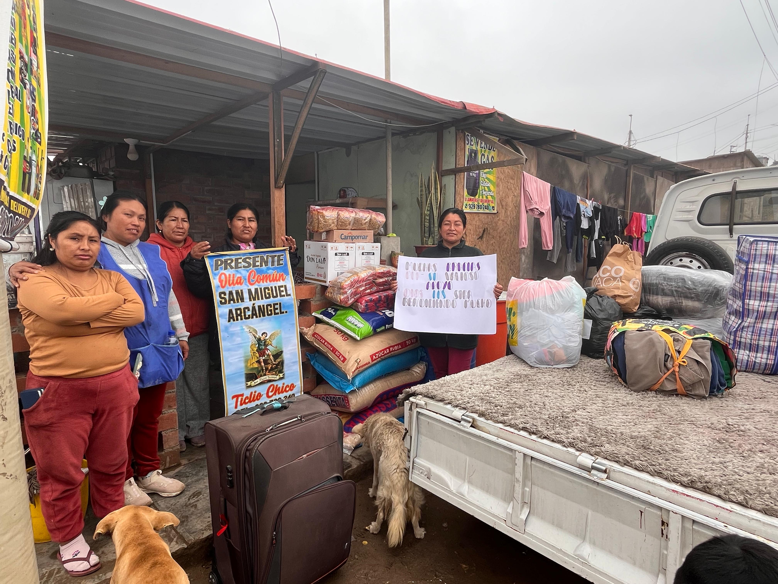 Group of women at a street market stall with bags of food and supplies. Two dogs, one golden and one black, are present. The women are standing behind tables with various items, including a sign that mentions Saint Michael Archangel. The scene is outdoors under a metal roof with a vehicle nearby.