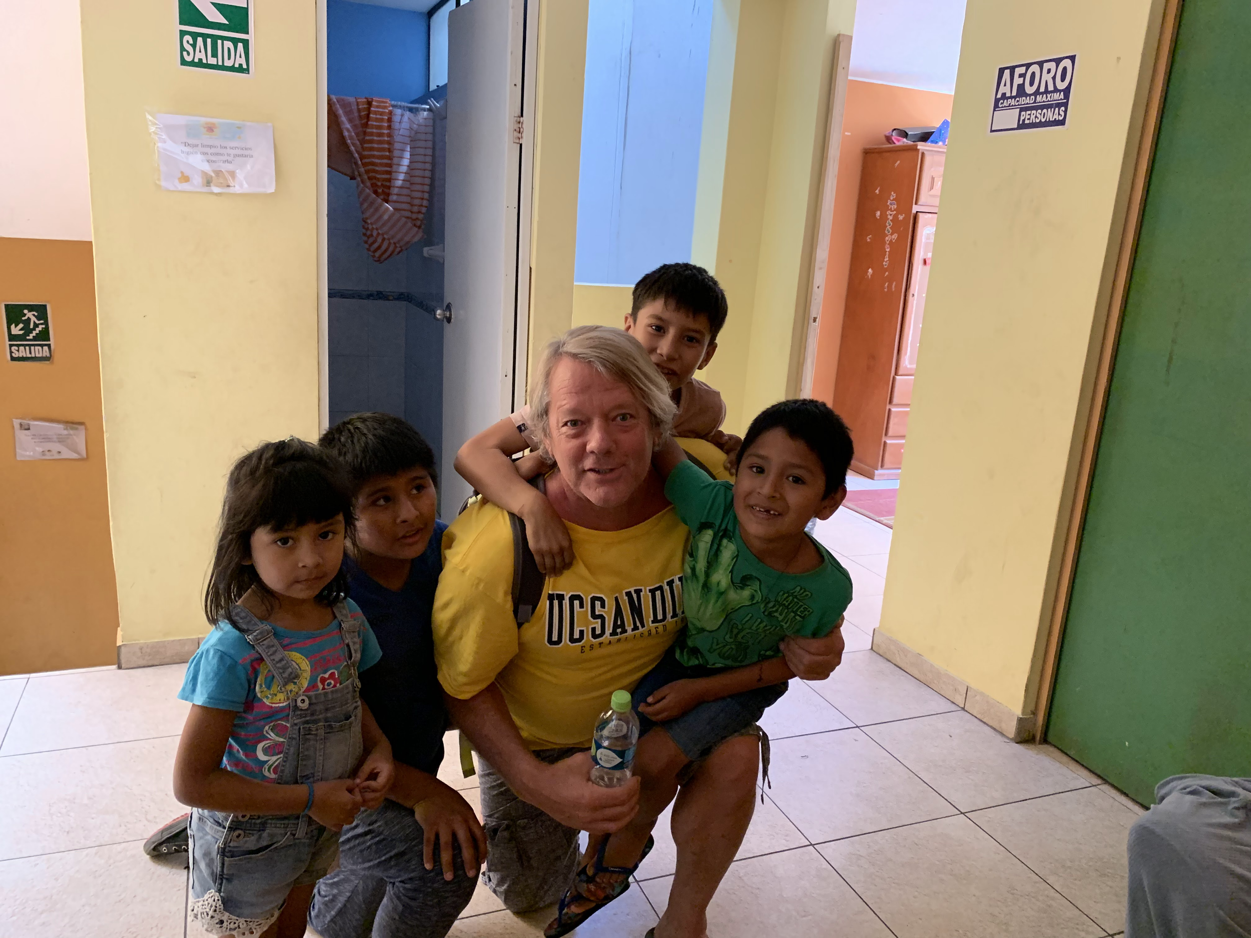 A group of children and an adult man posing together in a hallway with yellow walls. The children are smiling and standing close to the man, who is holding a water bottle. Part of the hallway with an open door and plants is visible in the background.