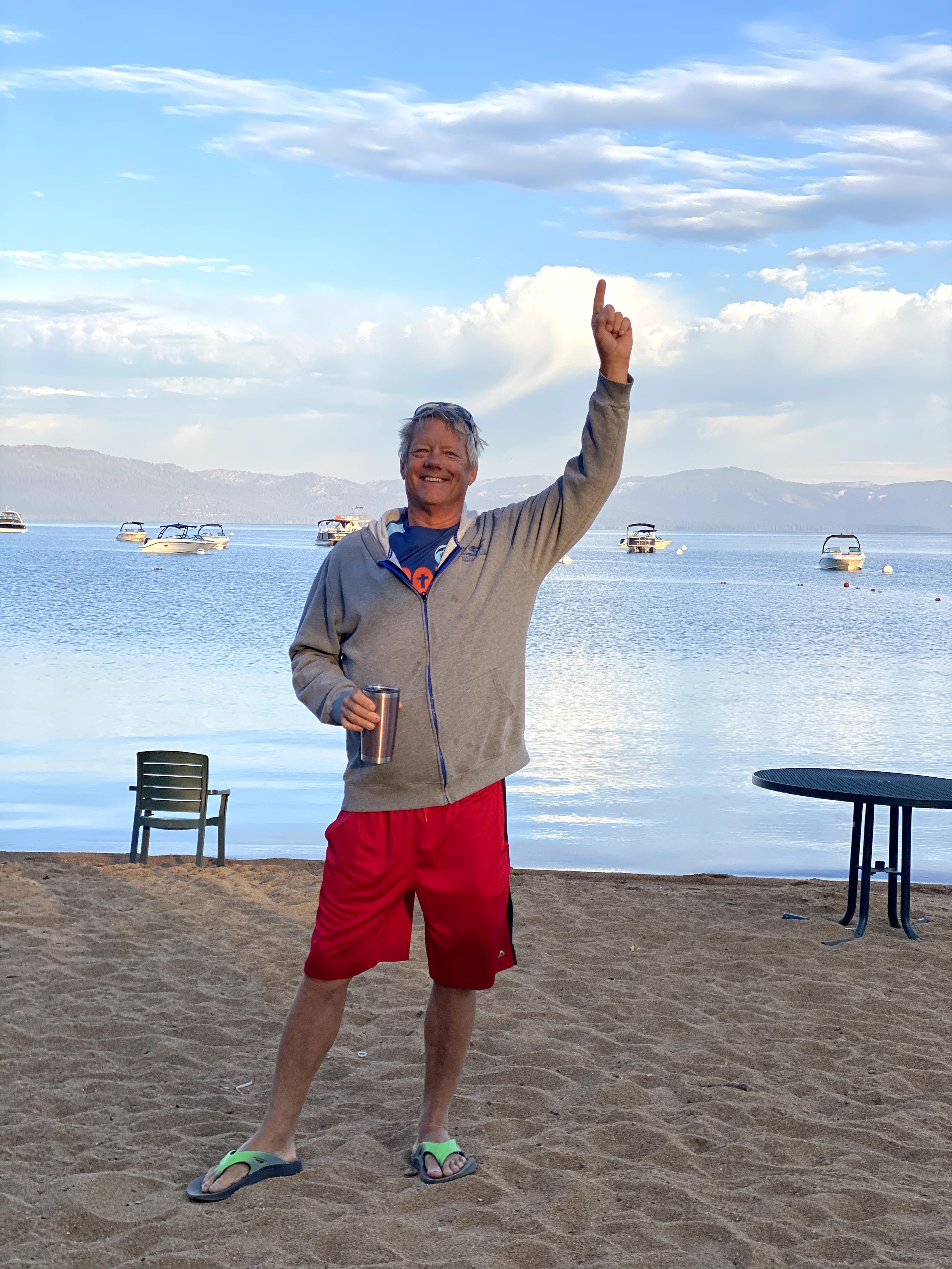 Smile of a middle-aged man, holding one arm up pointing to the sky and the other holding a tumbler, dressed in a light jacket and red shorts, standing on sandy beach with lake and mountains in the background under a partly cloudy sky.
