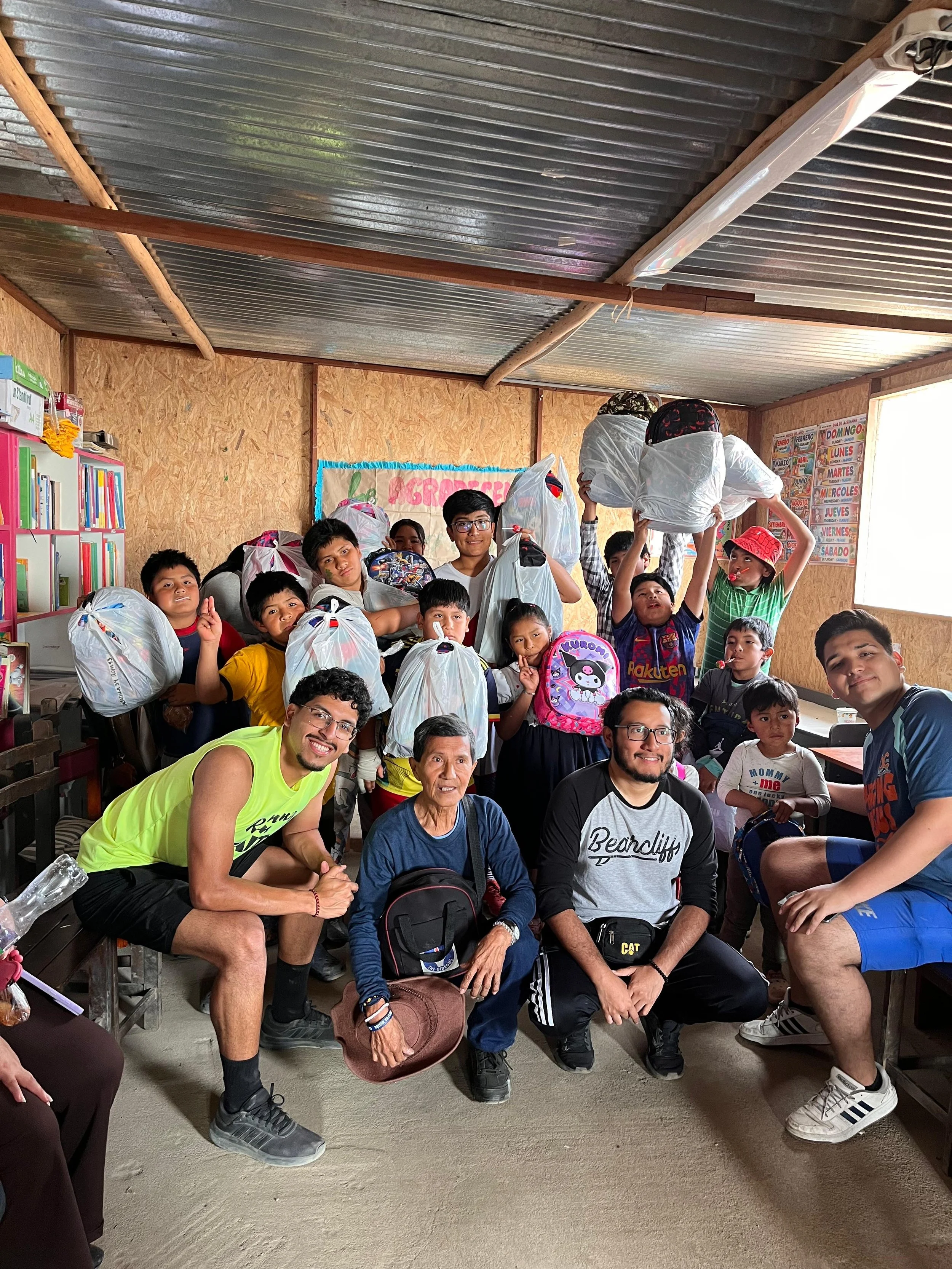 A group of children and adults in a room with plywood walls and a metal ceiling, some holding large plastic bags, smiling and posing for the camera.
