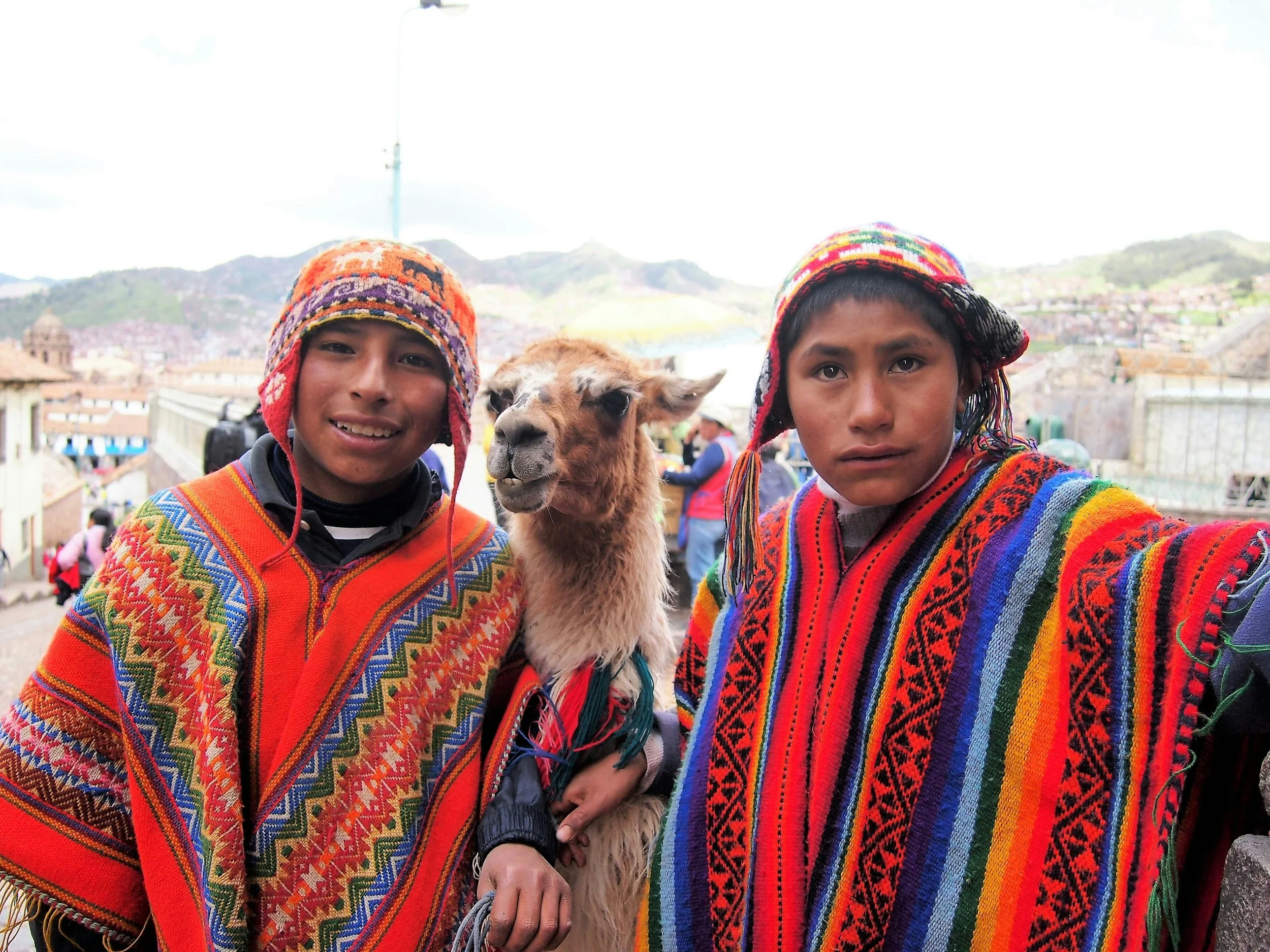 Two boys in colorful traditional woven clothing take a selfie with a llama. They are outdoors in a town with mountains in the background.
