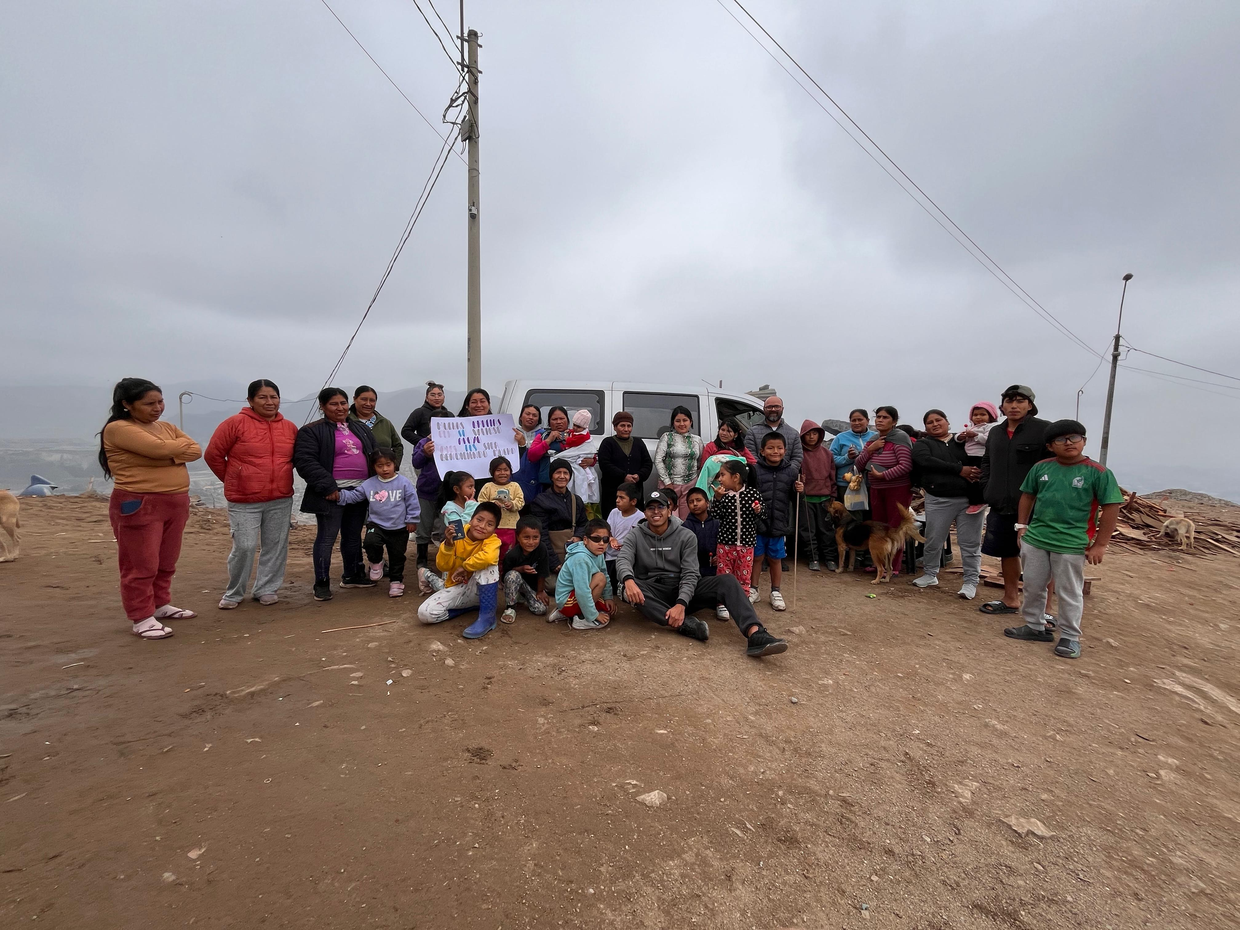 Group of people, including children, standing outdoors on dirt ground with a vehicle and power lines in the background on a cloudy day.