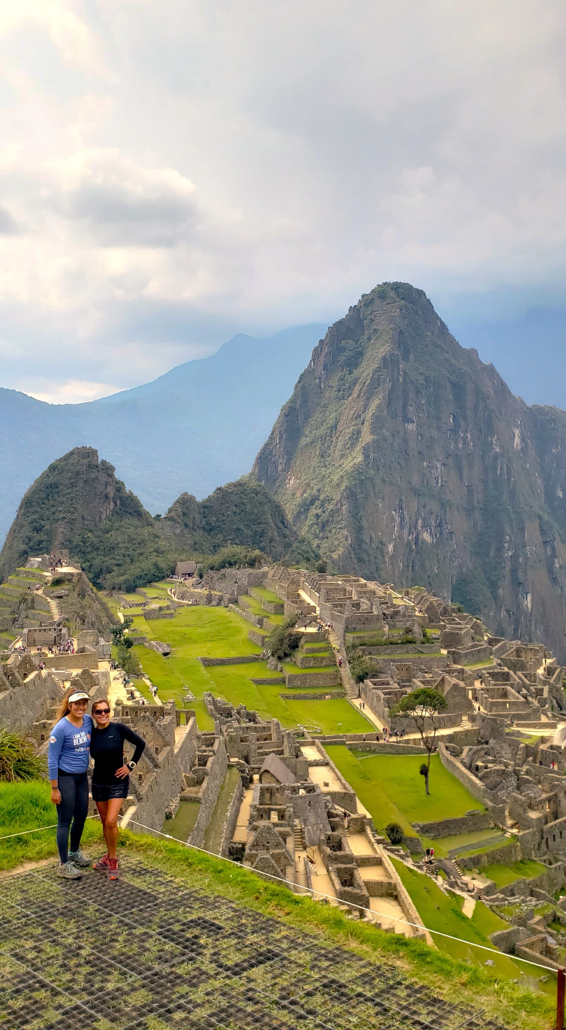 Two women standing and smiling in front of Machu Picchu with ancient stone ruins, terraces, and the iconic mountain peak in the background.