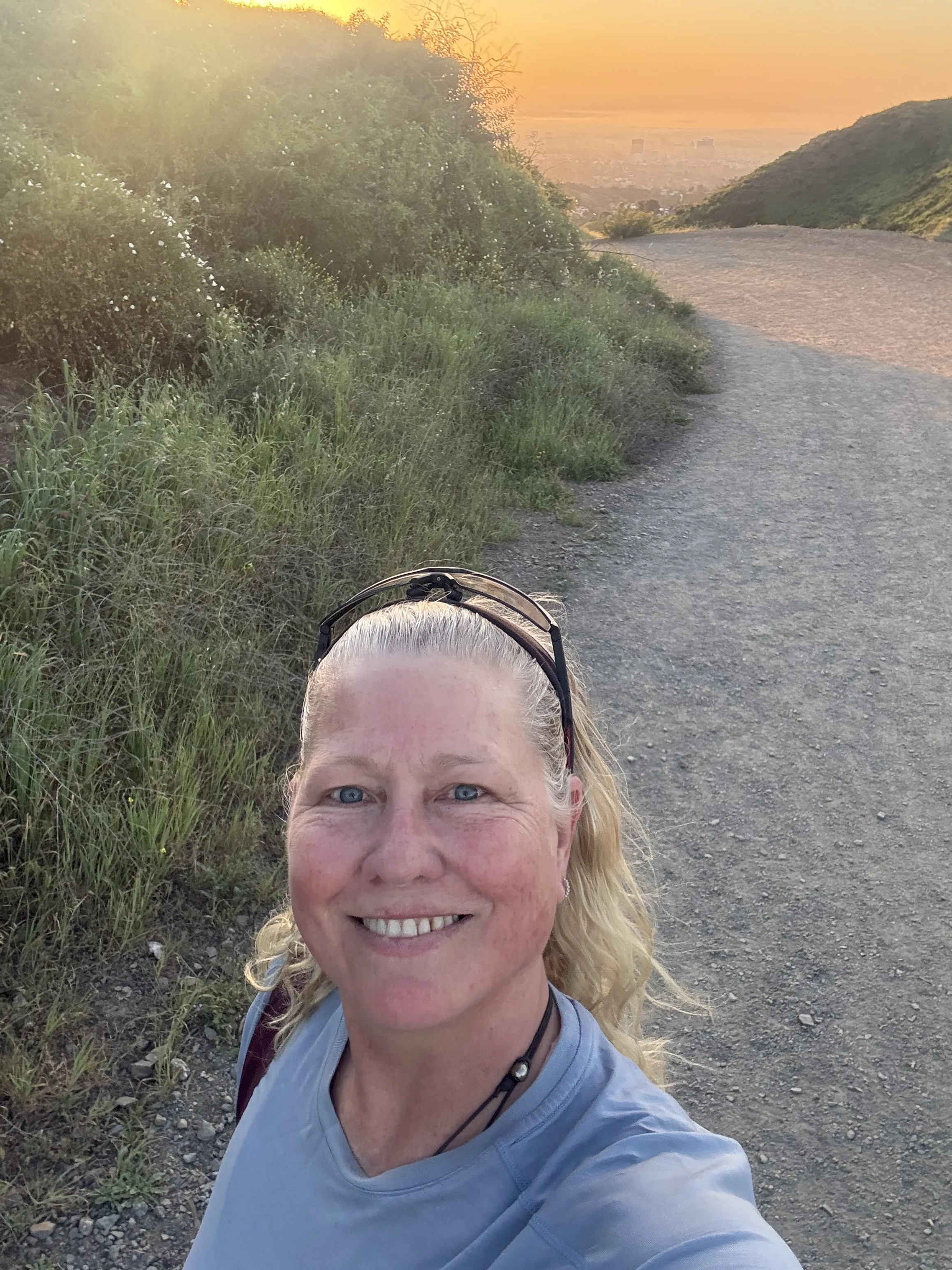 A smiling woman with blonde hair, wearing a light blue shirt and sunglasses on her head, takes a selfie on a dirt trail near a hillside at sunset.