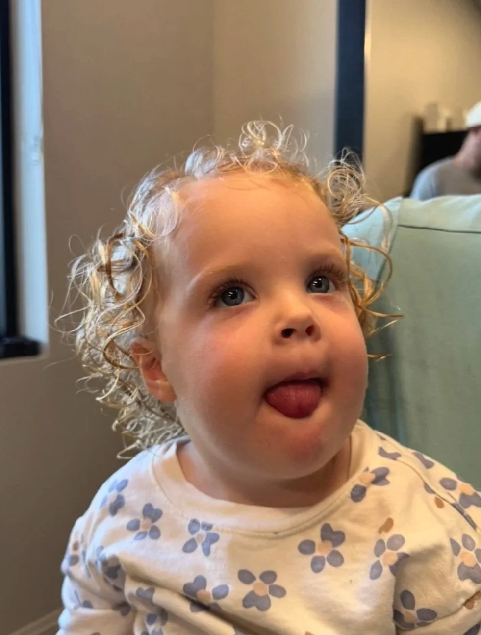 A young child with curly blonde hair sticking out their tongue, sitting on a person's lap in a room with a window and a man wearing a baseball cap in the background.