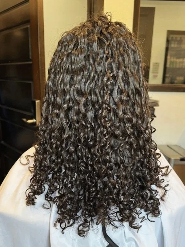 Back view of a person with shoulder-length, dark brown, tight curly hair, sitting in a salon chair wearing a white cape.