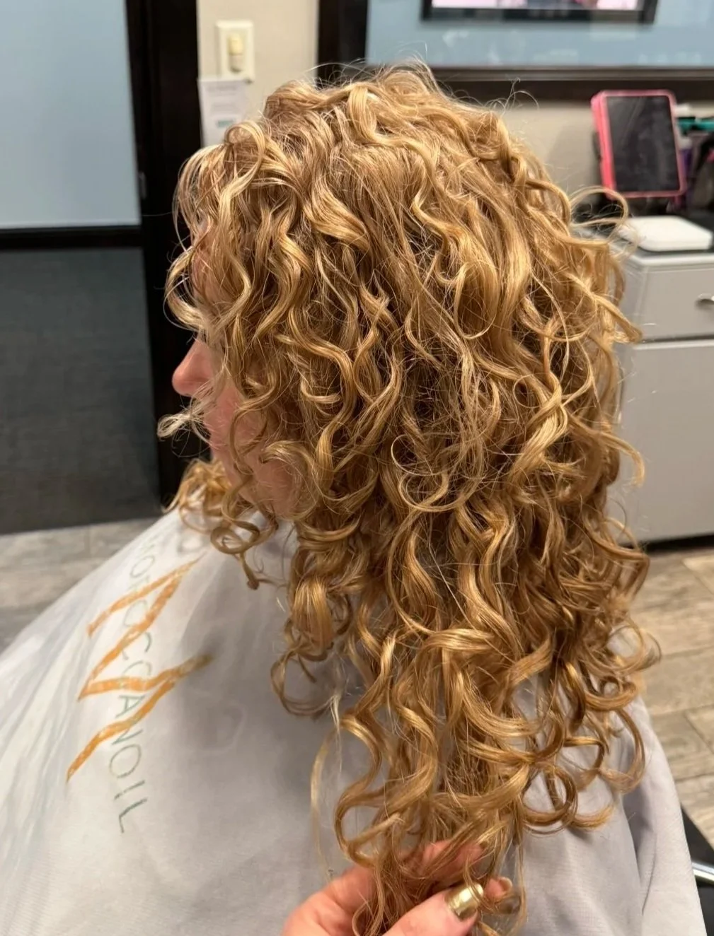 Close-up of a woman with long, curly, blonde hair at a hair salon, wearing a salon cape, while a hand gently holds a section of her curly hair.